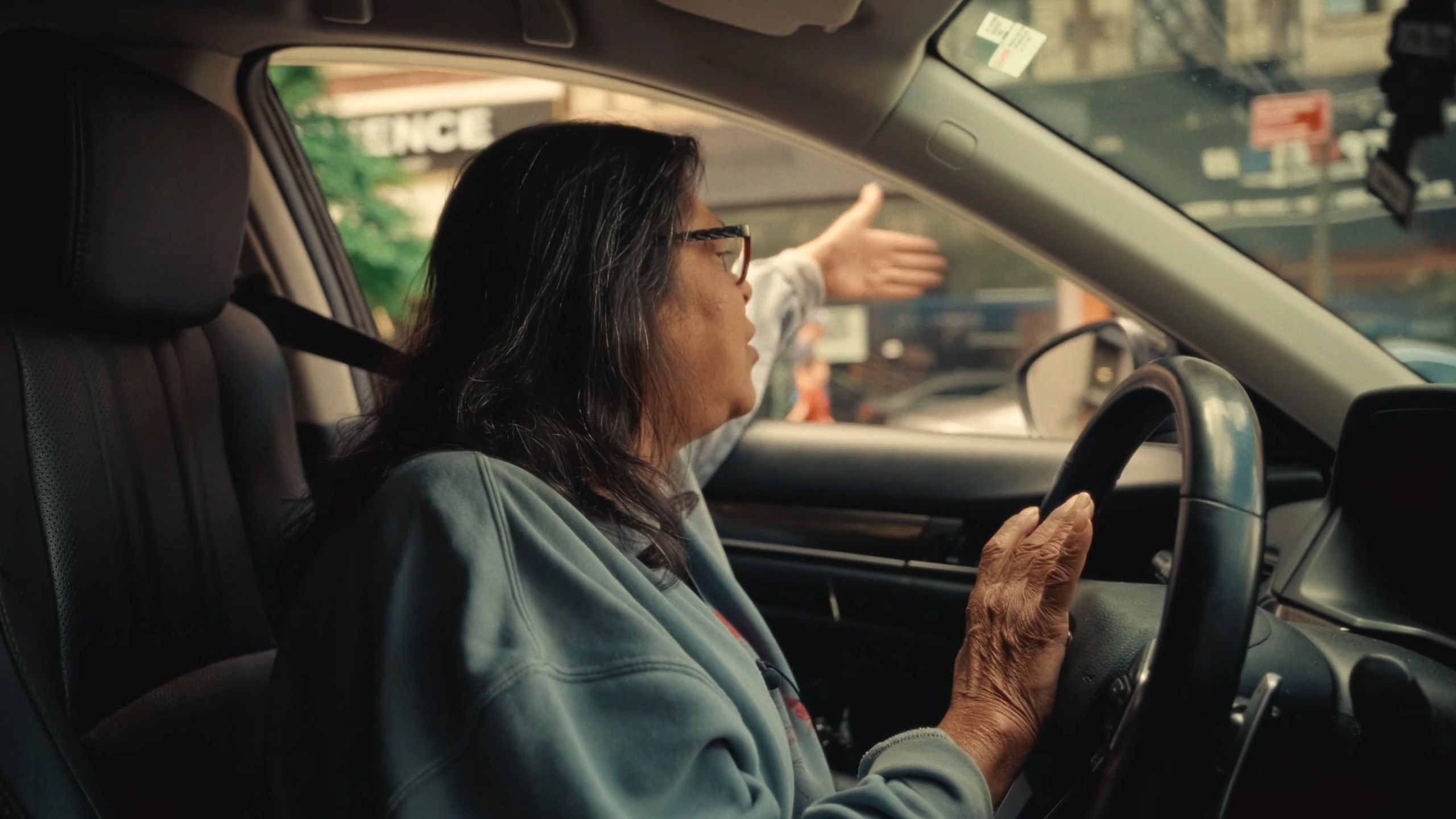 Photo of a person with long hair and glasses driving a car gesturing to someone outside through an open window.