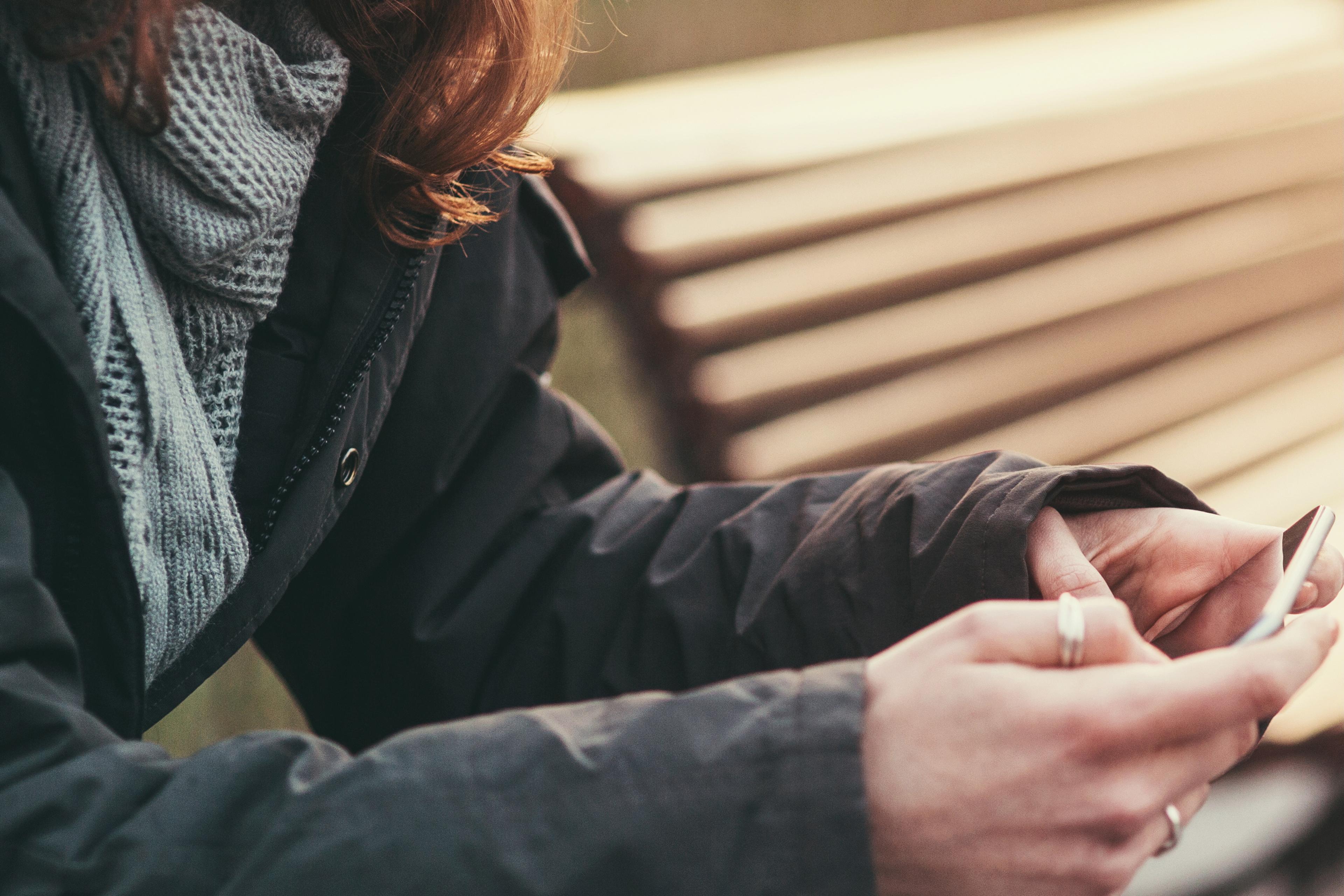Photo of a person in a coat using a smartphone outdoors, sitting on a bench with focus on hands and partial face in view.