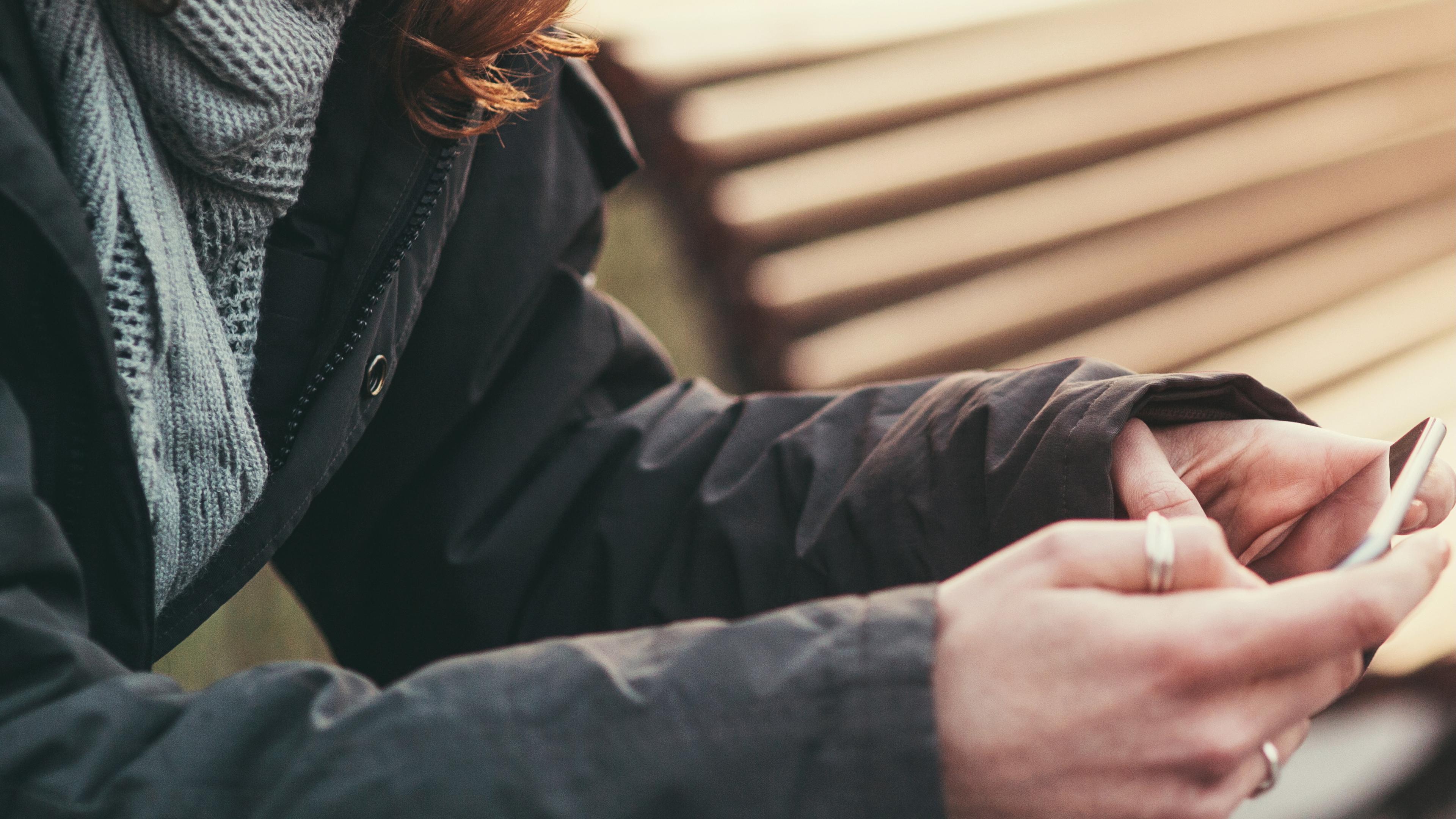 Photo of a person in a coat using a smartphone outdoors, sitting on a bench with focus on hands and partial face in view.