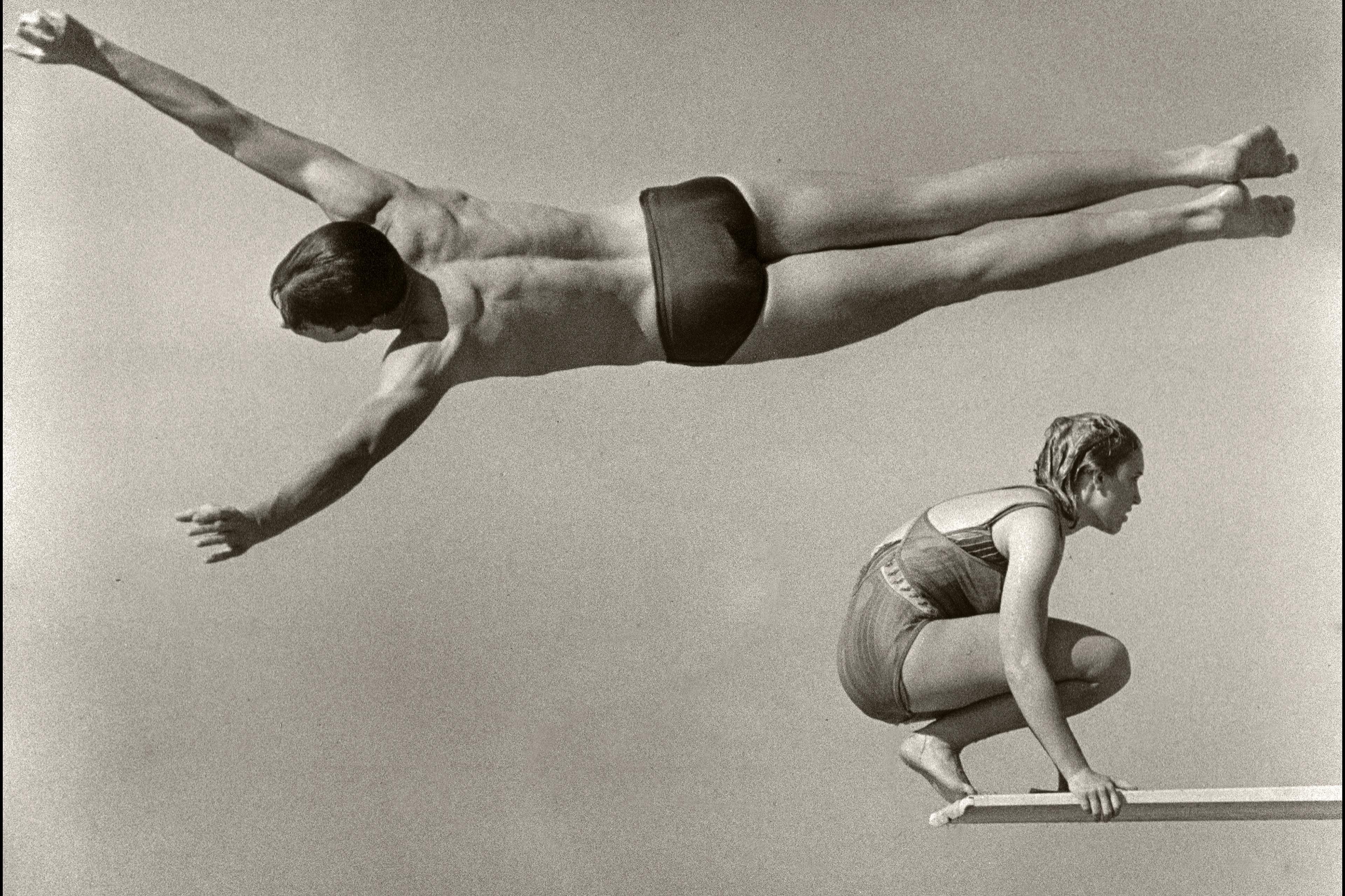 Black and white photo of a diver mid-flight parallel to a crouched swimmer on a diving board against a plain background.