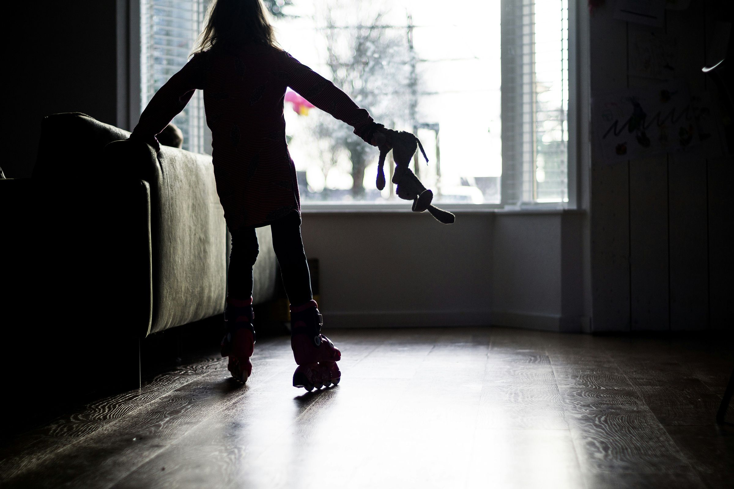 Silhouette photo of a child in roller skates holding a toy bunny, standing by a sofa near a bright window.