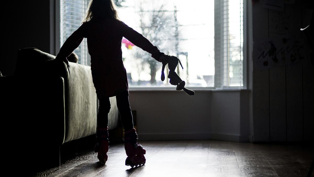 Silhouette photo of a child in roller skates holding a toy bunny, standing by a sofa near a bright window.