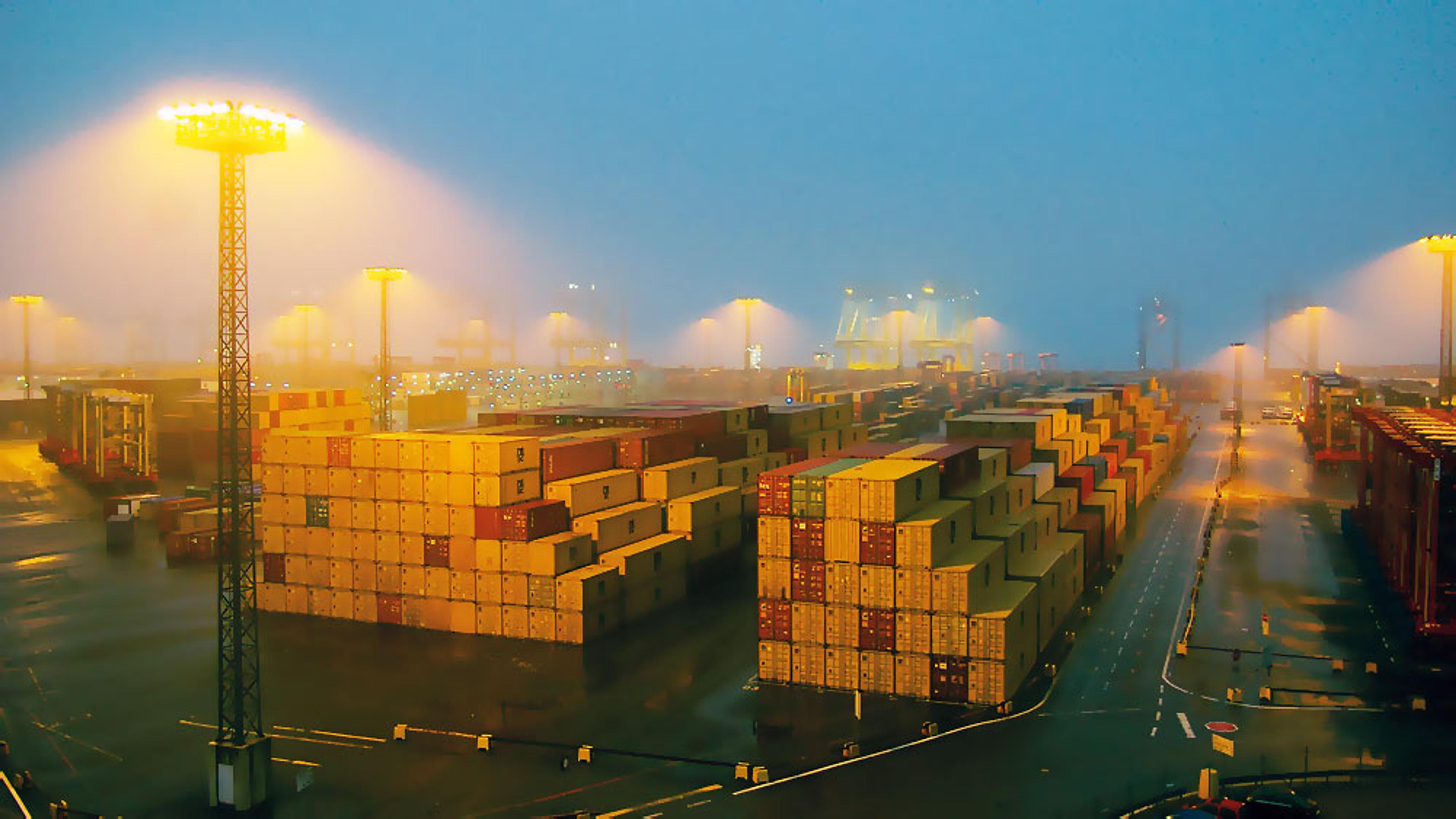 A cargo port at dusk with stacked shipping containers and bright overhead lights illuminating the area and causing a light haze.
