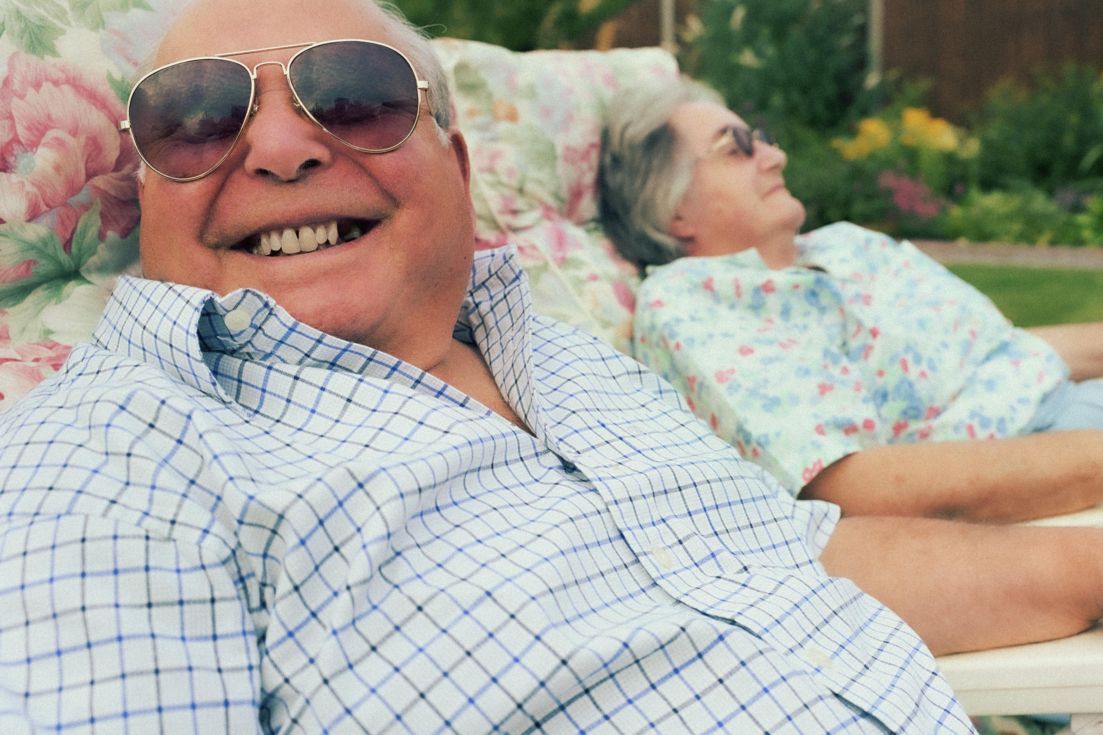 An elderly man smiling broadly with sunglasses on, reclining on a floral lounge chair beside an elderly woman outdoors.
