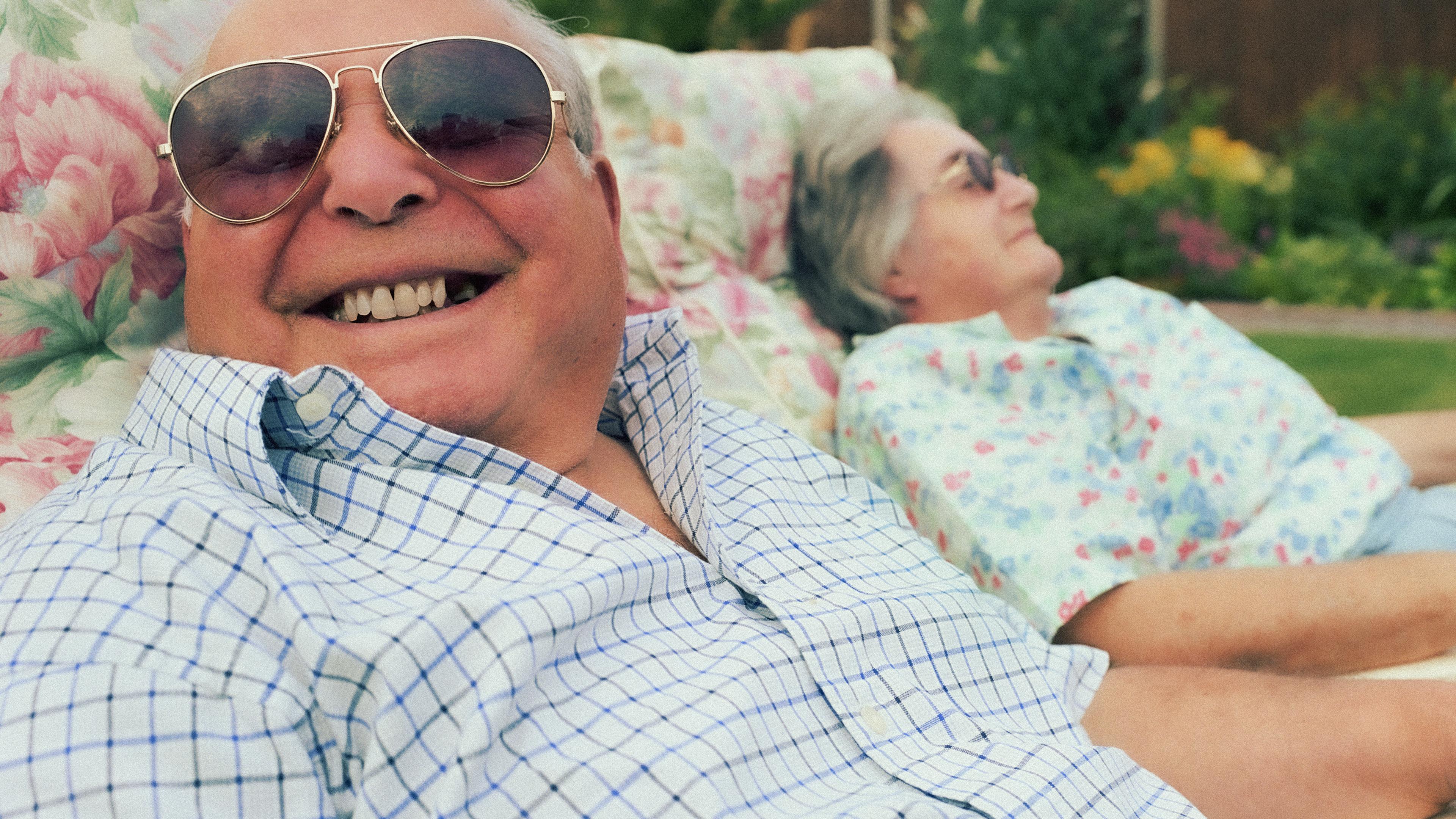 An elderly man smiling broadly with sunglasses on, reclining on a floral lounge chair beside an elderly woman outdoors.