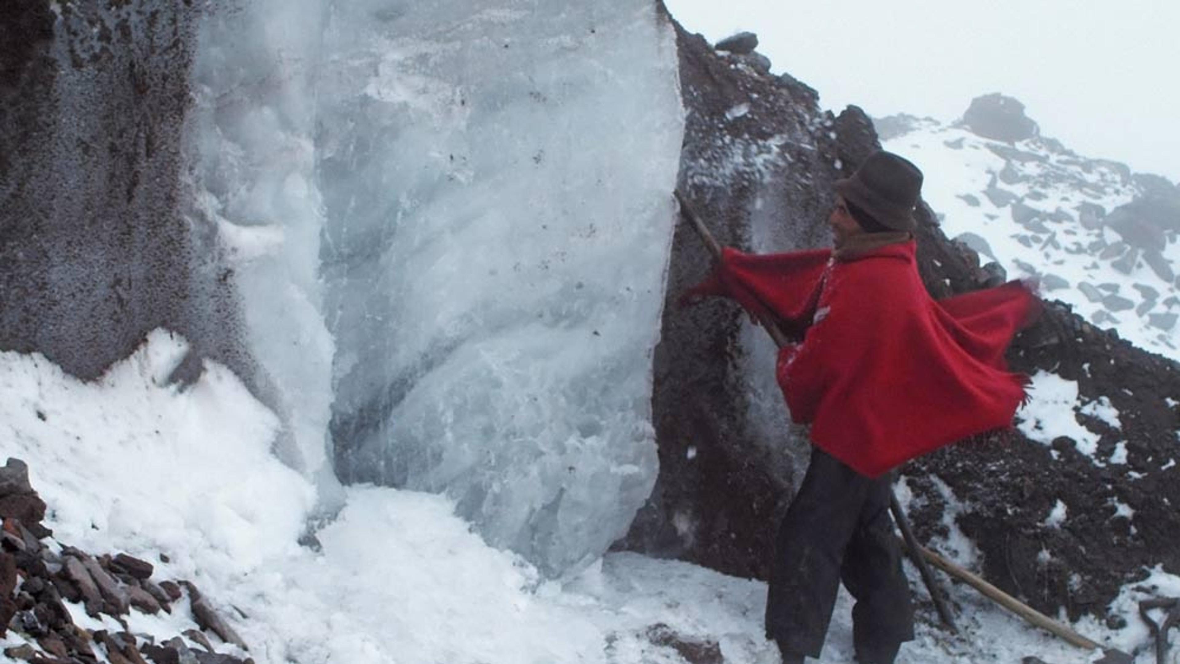 A person in a red poncho and hat using a tool to extract ice from a snowy, rocky mountainside.