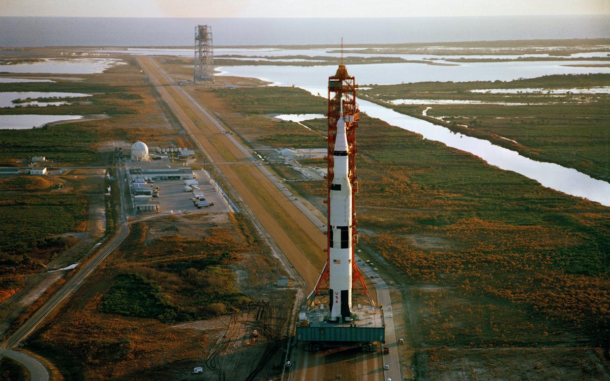 Aerial photo of a large rocket on a mobile platform at a space launch facility, surrounded by water and greenery, leading to the ocean.