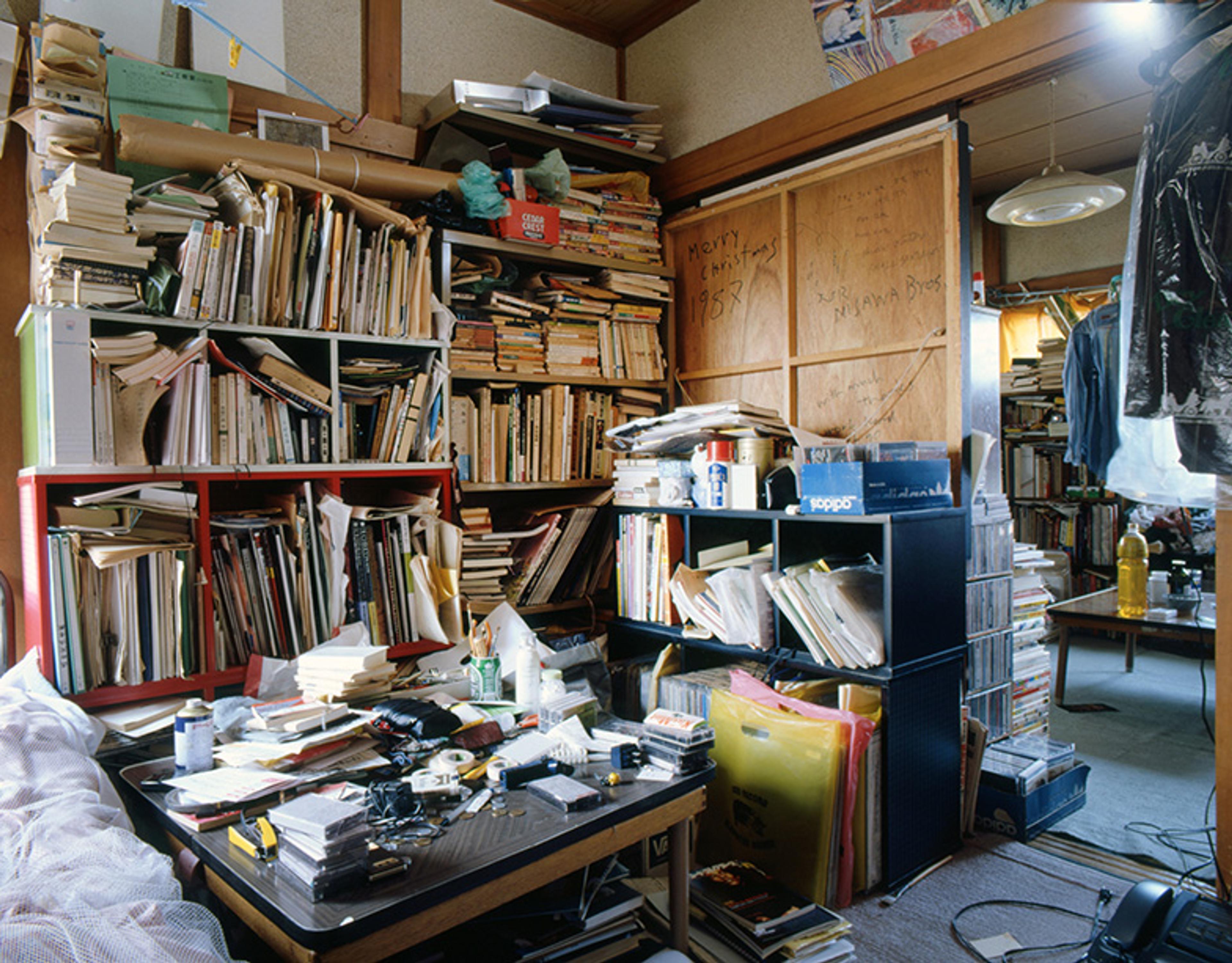 A cluttered room with overflowing bookshelves, a disorganised desk covered with items, and various objects strewn about.