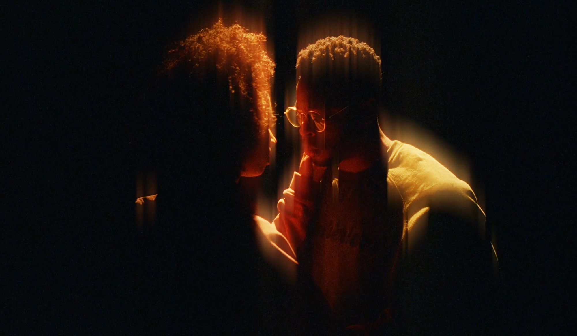 Two people standing closely in a dark setting, illuminated softly from above, creating a warm, intimate glow around them.