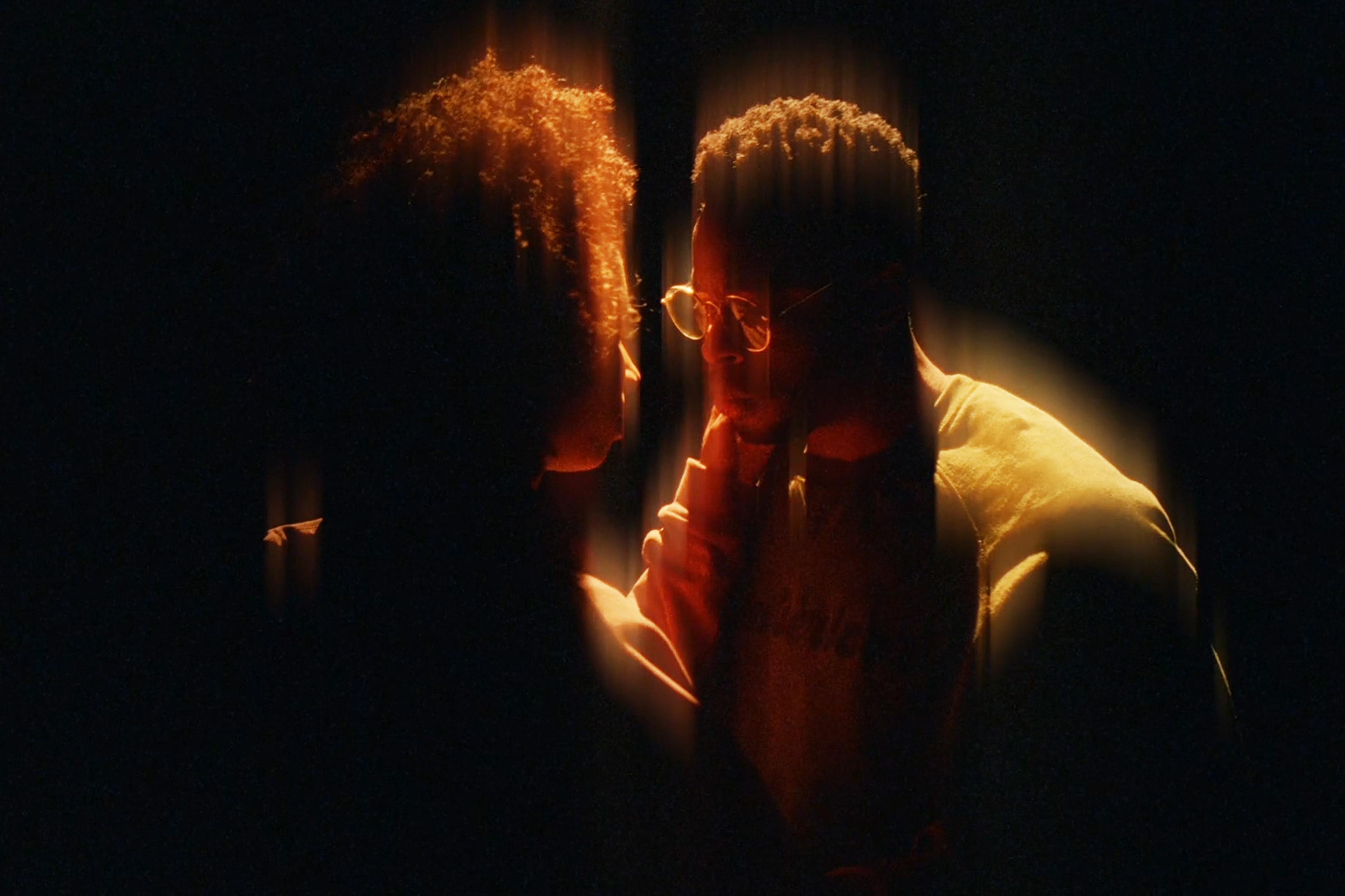 Two people standing closely in a dark setting, illuminated softly from above, creating a warm, intimate glow around them.