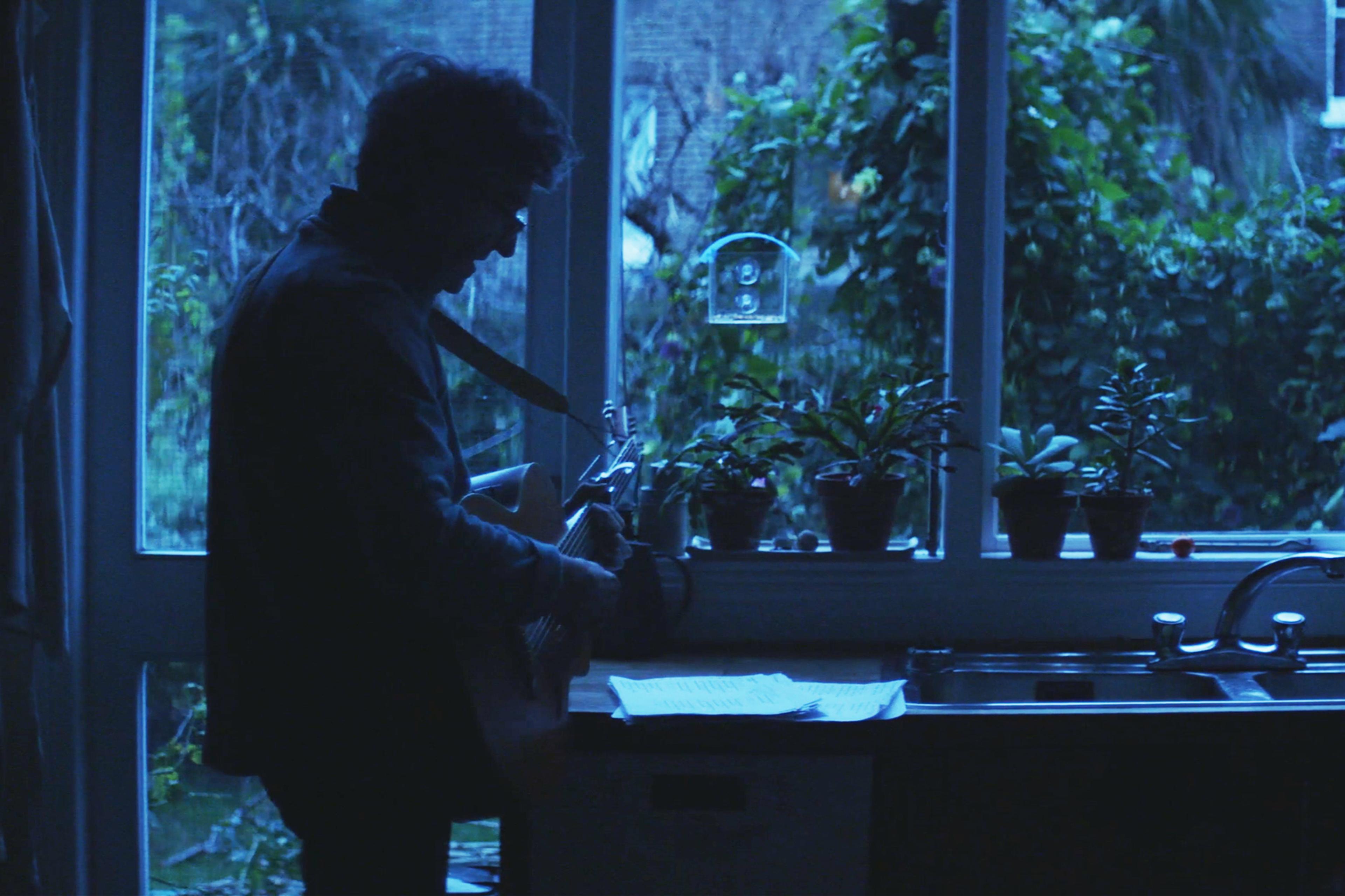 Silhouetted man playing guitar in a dimly lit room near a window with plants on the sill and music sheets on the counter.