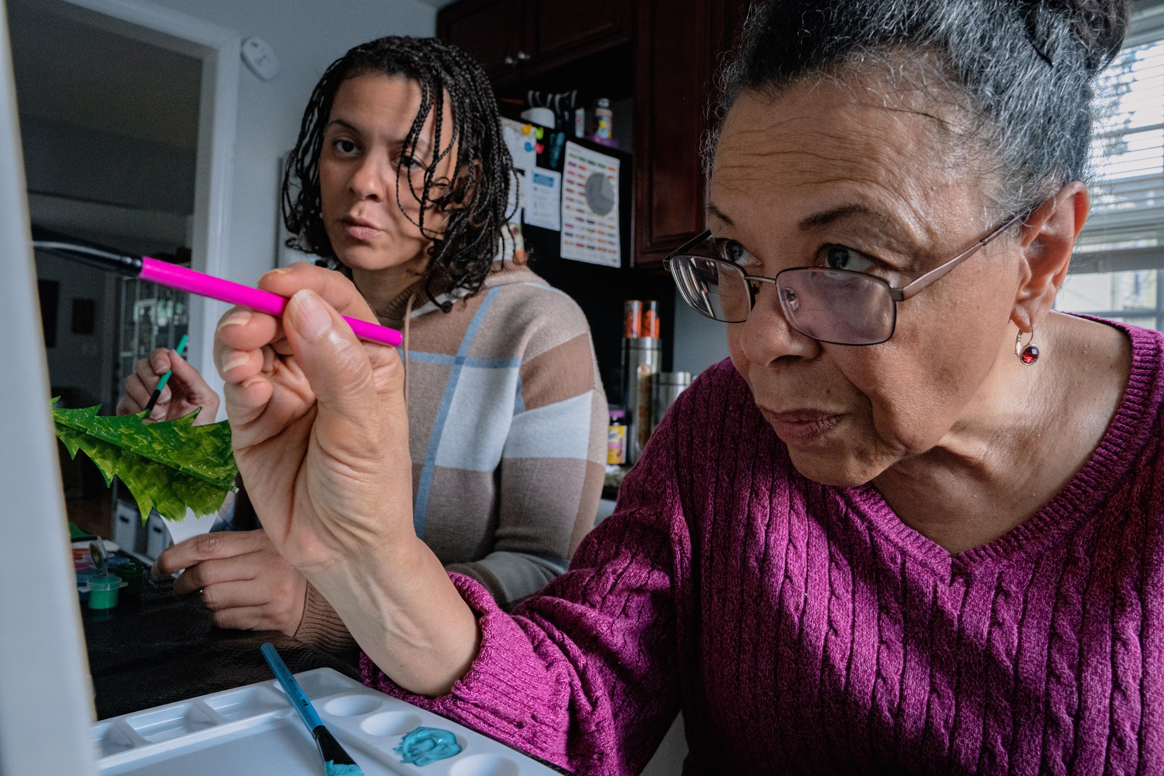 Photo of two women painting at a table, focusing on one using a pink brush.