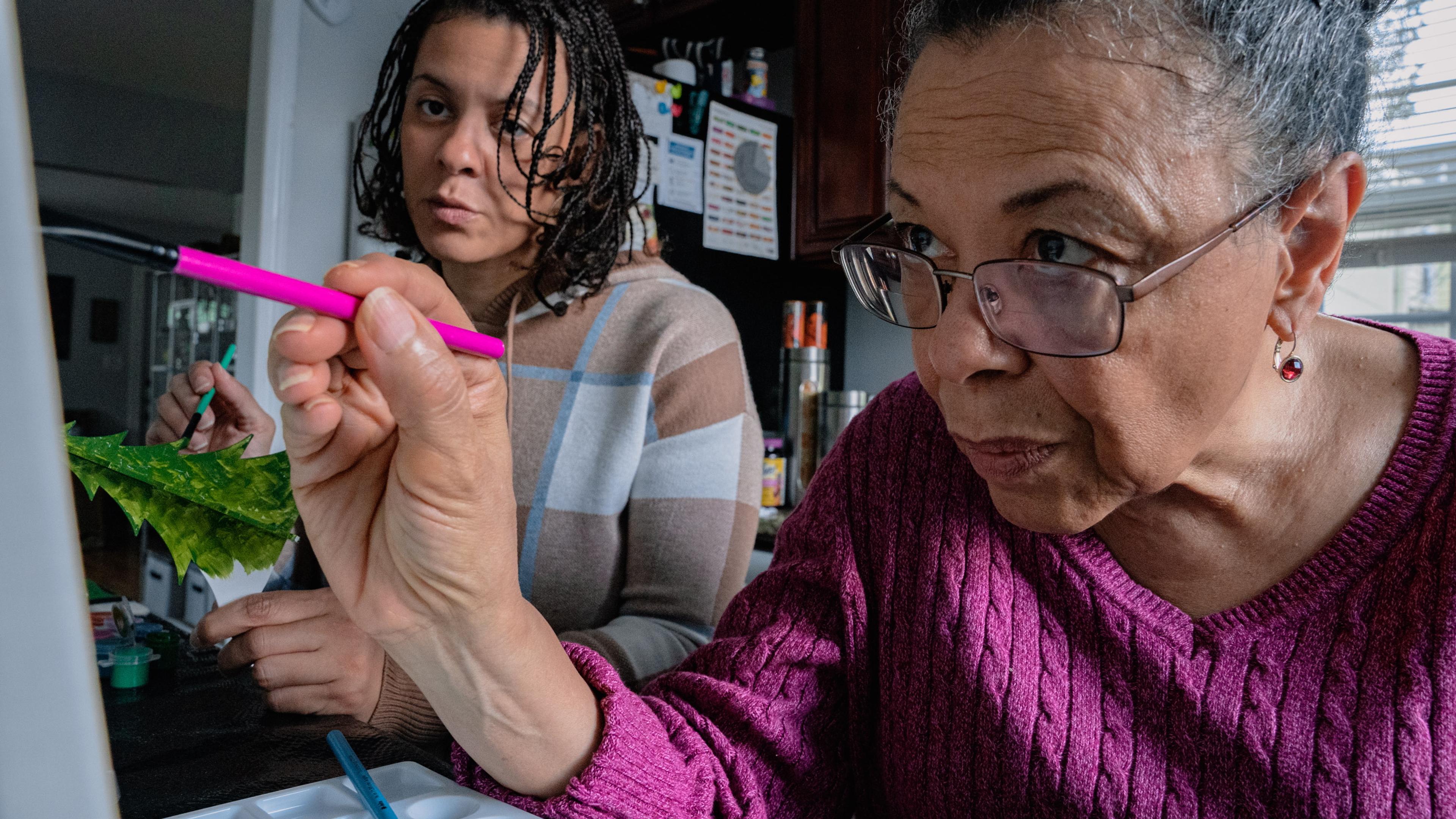 Photo of two women painting at a table, focusing on one using a pink brush.
