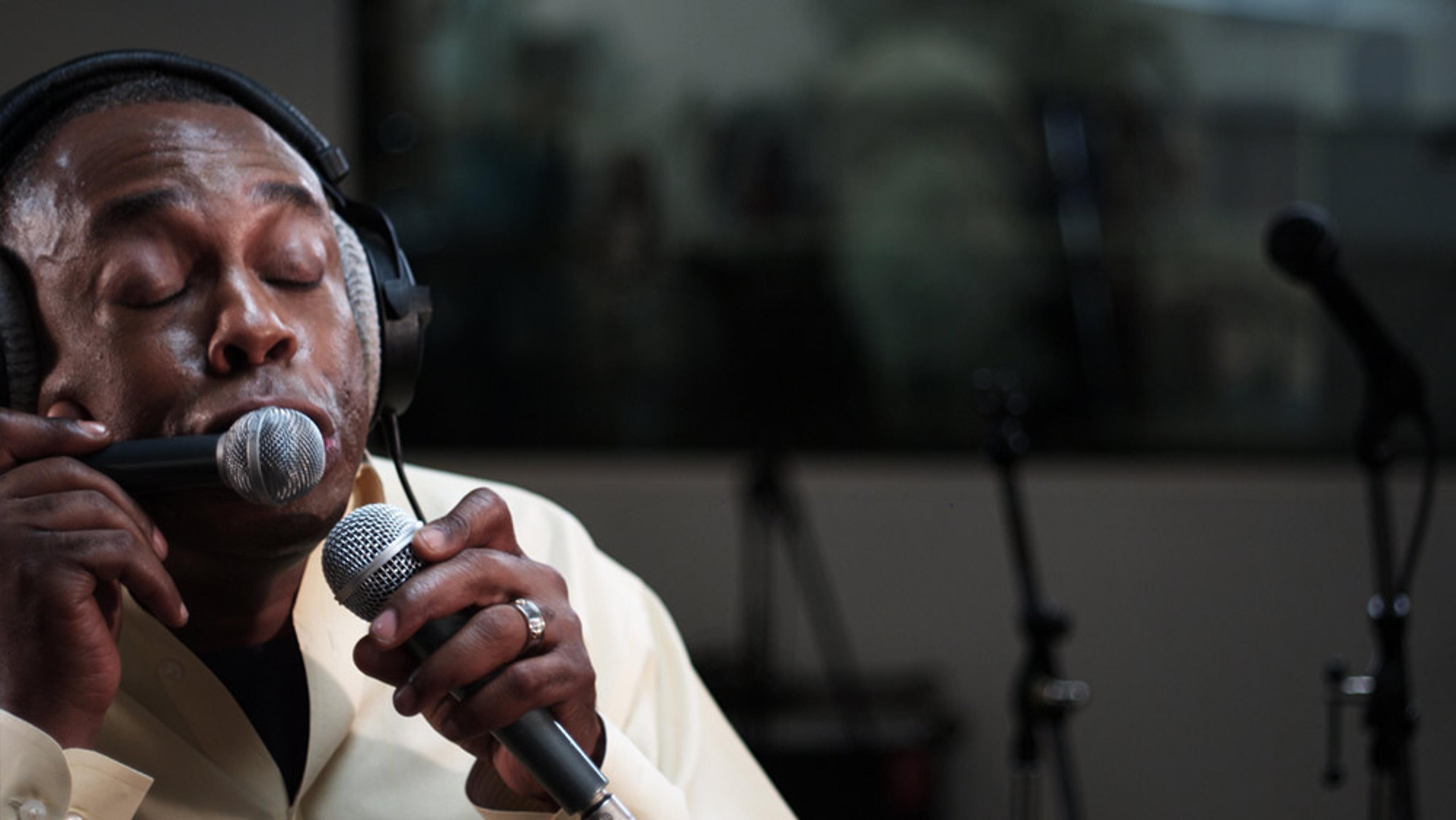 A man wearing headphones singing into a microphone with his eyes closed in a recording studio, holding another object to his mouth.