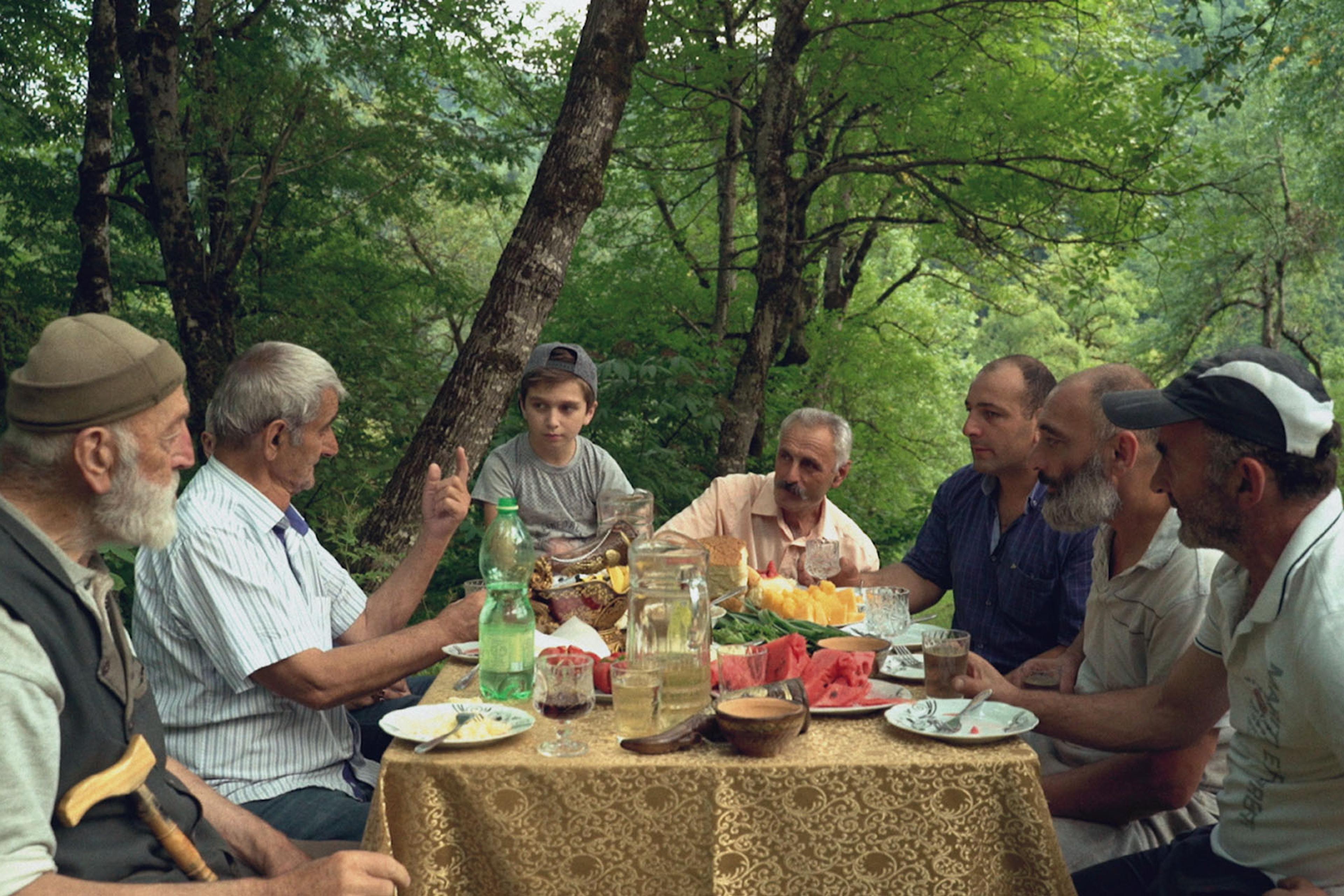 Group of men and a boy dining at an outdoor table in a forest, with various foods and drinks on a lace tablecloth.