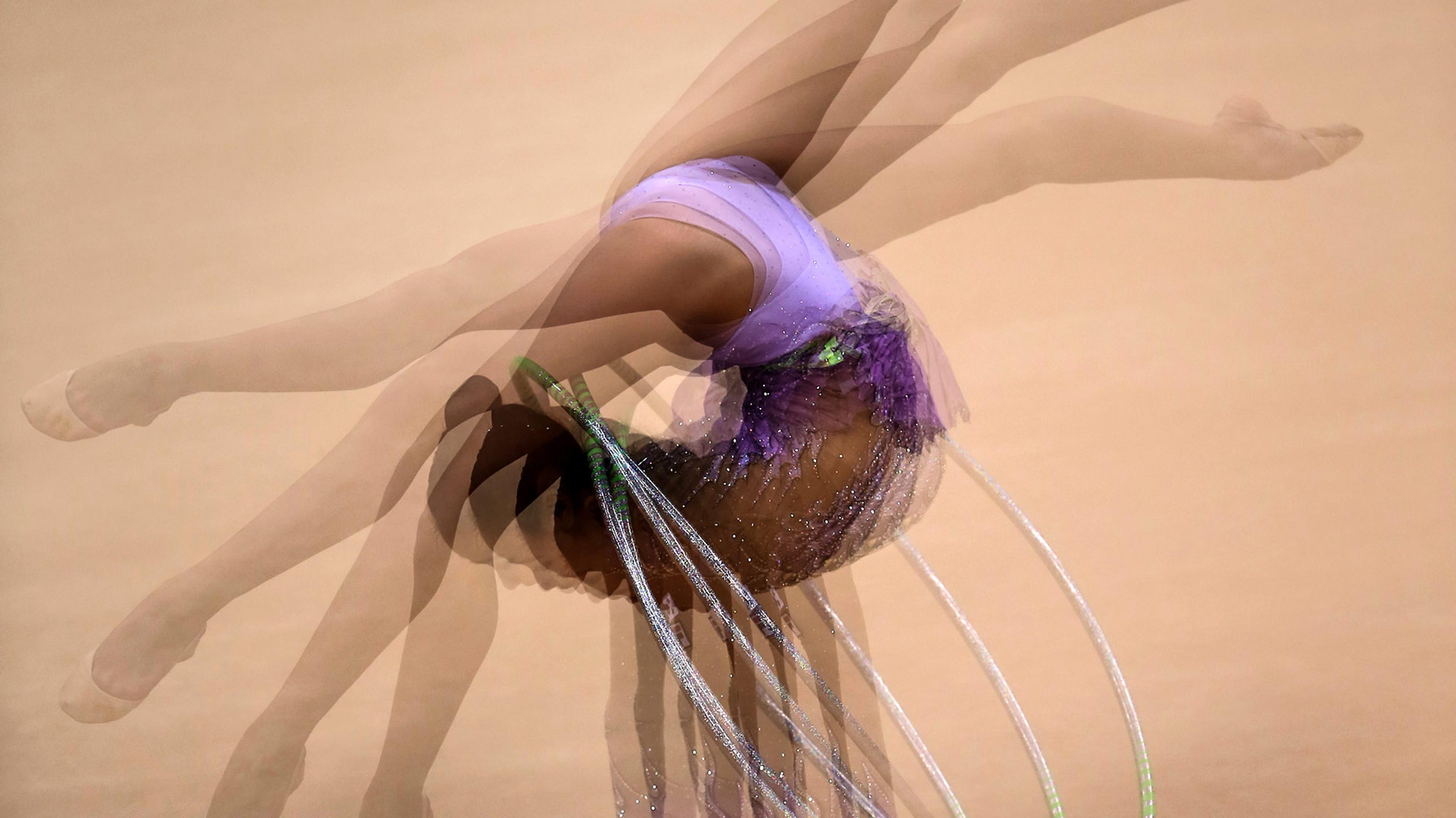 A gymnast in a purple leotard mid-performance, captured with a motion blur effect, showing multiple overlapping images of her movement.