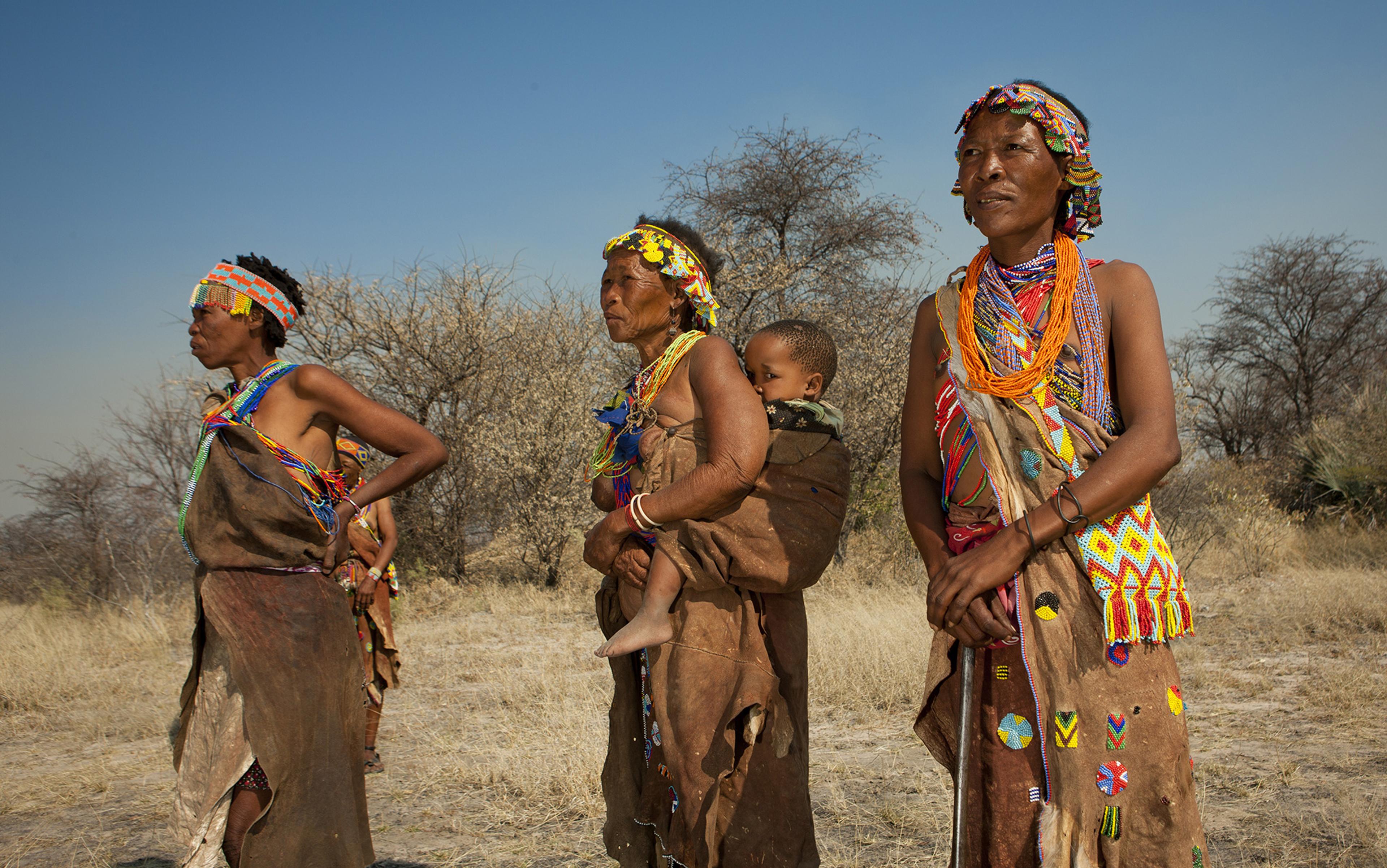 Three women in traditional attire stand outdoors in a dry landscape. One person carries a child on their back while another holds a walking stick.