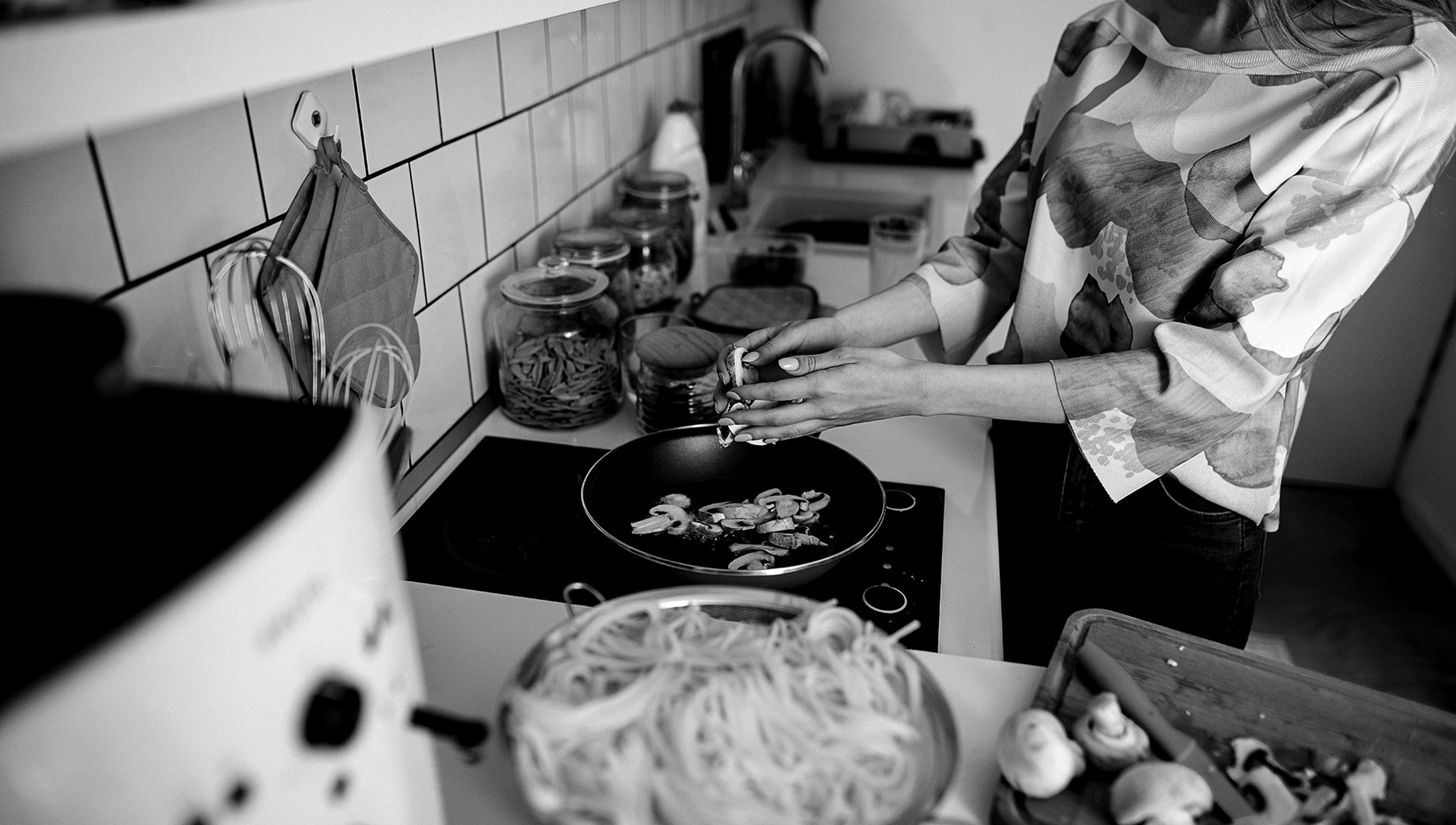 A person cooks mushrooms in a pan on a stove with pasta in a colander and various ingredients spread on the kitchen counter.