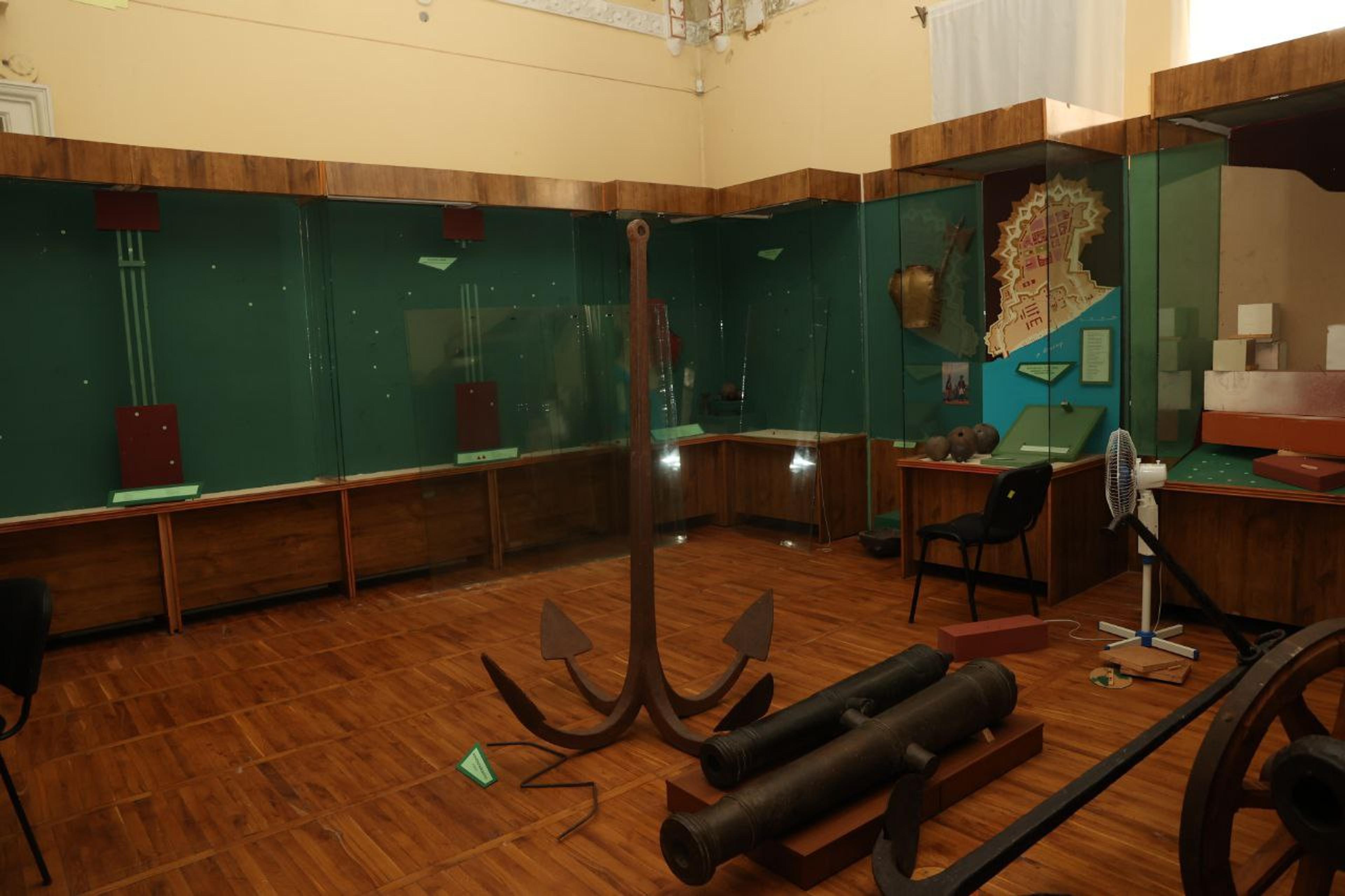 A museum exhibit room with an anchor, cannons and other historical artefacts displayed on wooden floors and glass cases.