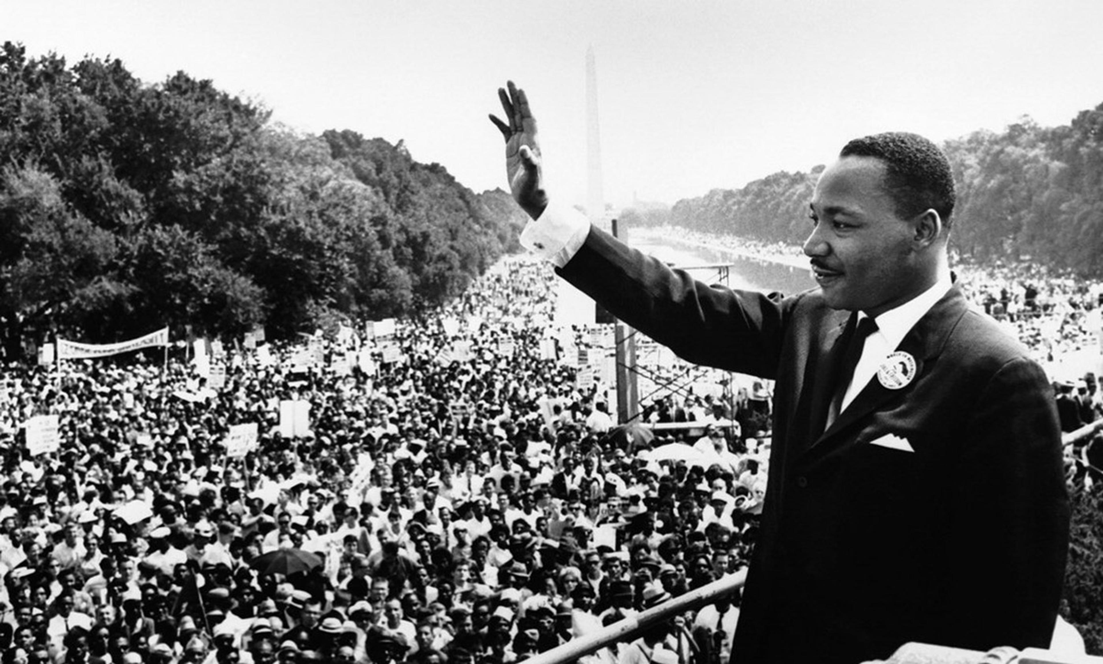 Vintage photo of Martin Luther King Jr in a suit and tie waving to a large crowd during a civil rights march with trees and a monument in the background.
