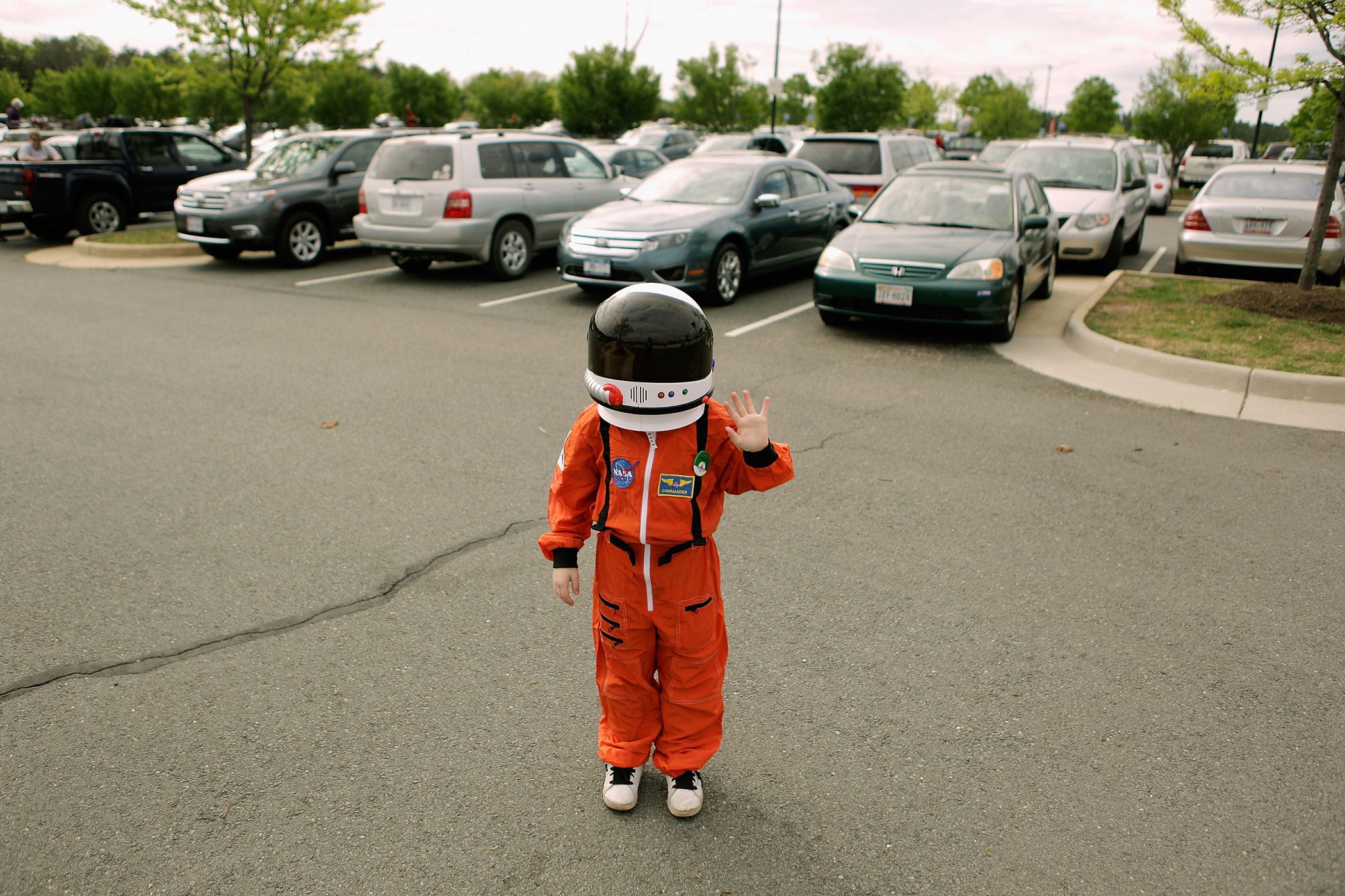 A child in an orange astronaut suit waving in a car park with cars and trees in the background.