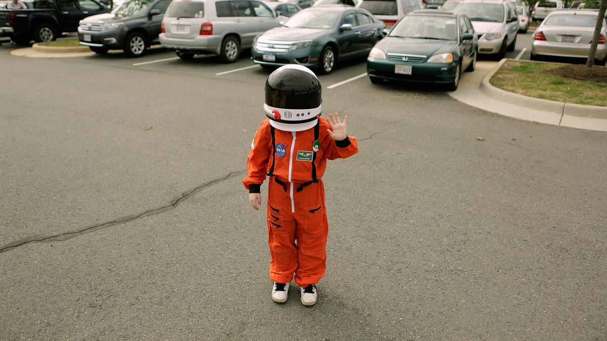 A child in an orange astronaut suit waving in a car park with cars and trees in the background.