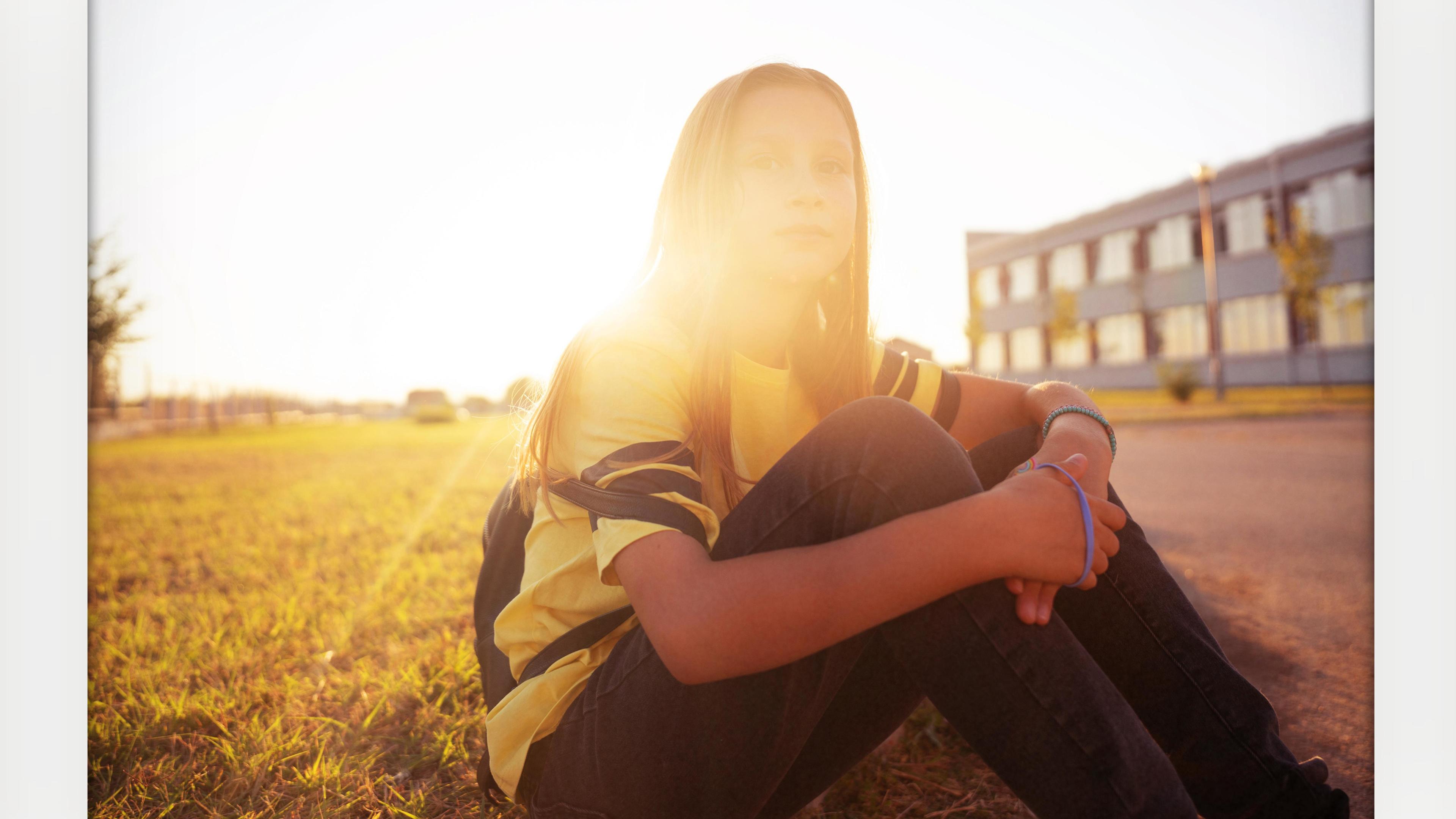 A person sitting in sunlight on grass by a building, wearing a yellow top and jeans, with soft lens flare.