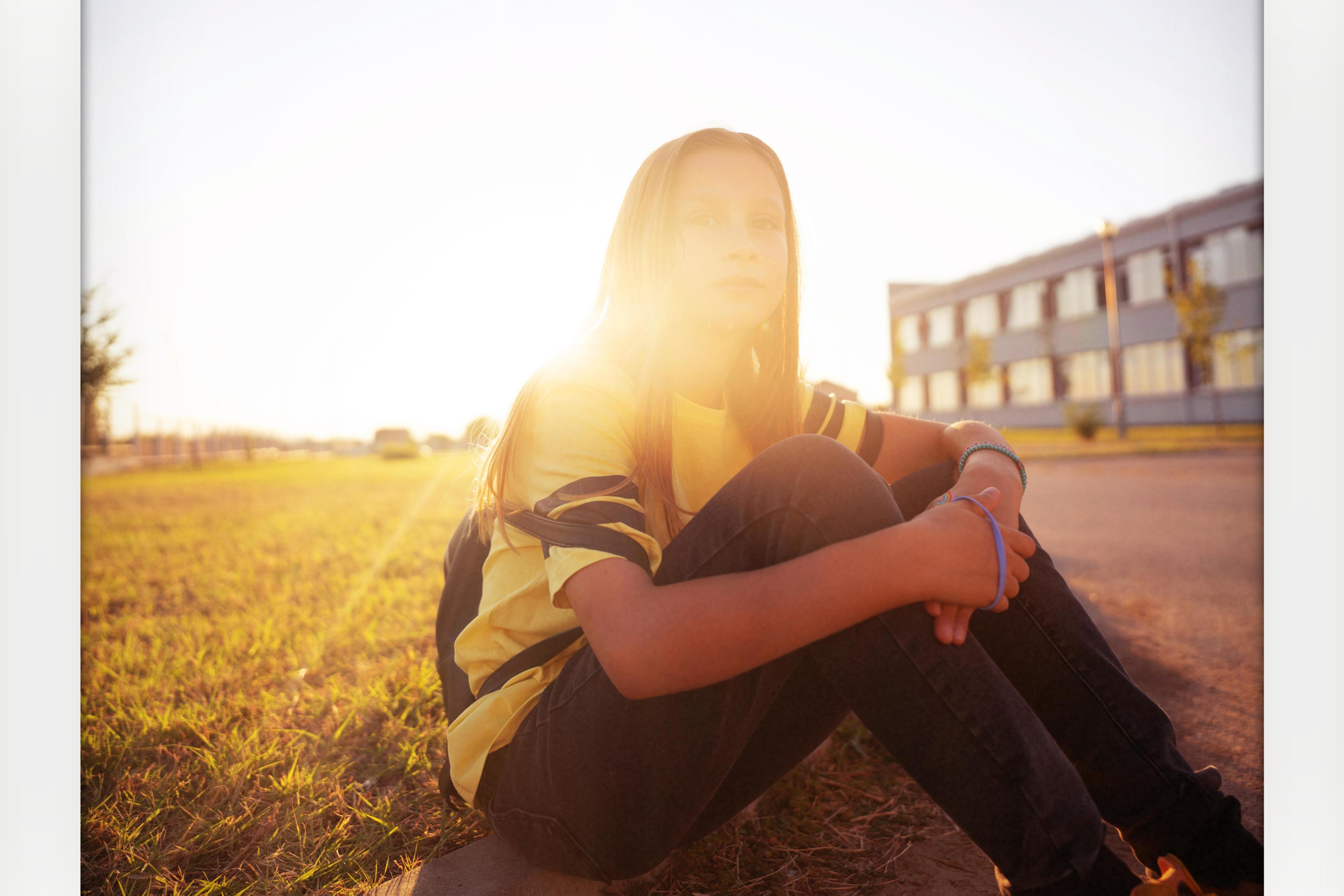 A person sitting in sunlight on grass by a building, wearing a yellow top and jeans, with soft lens flare.