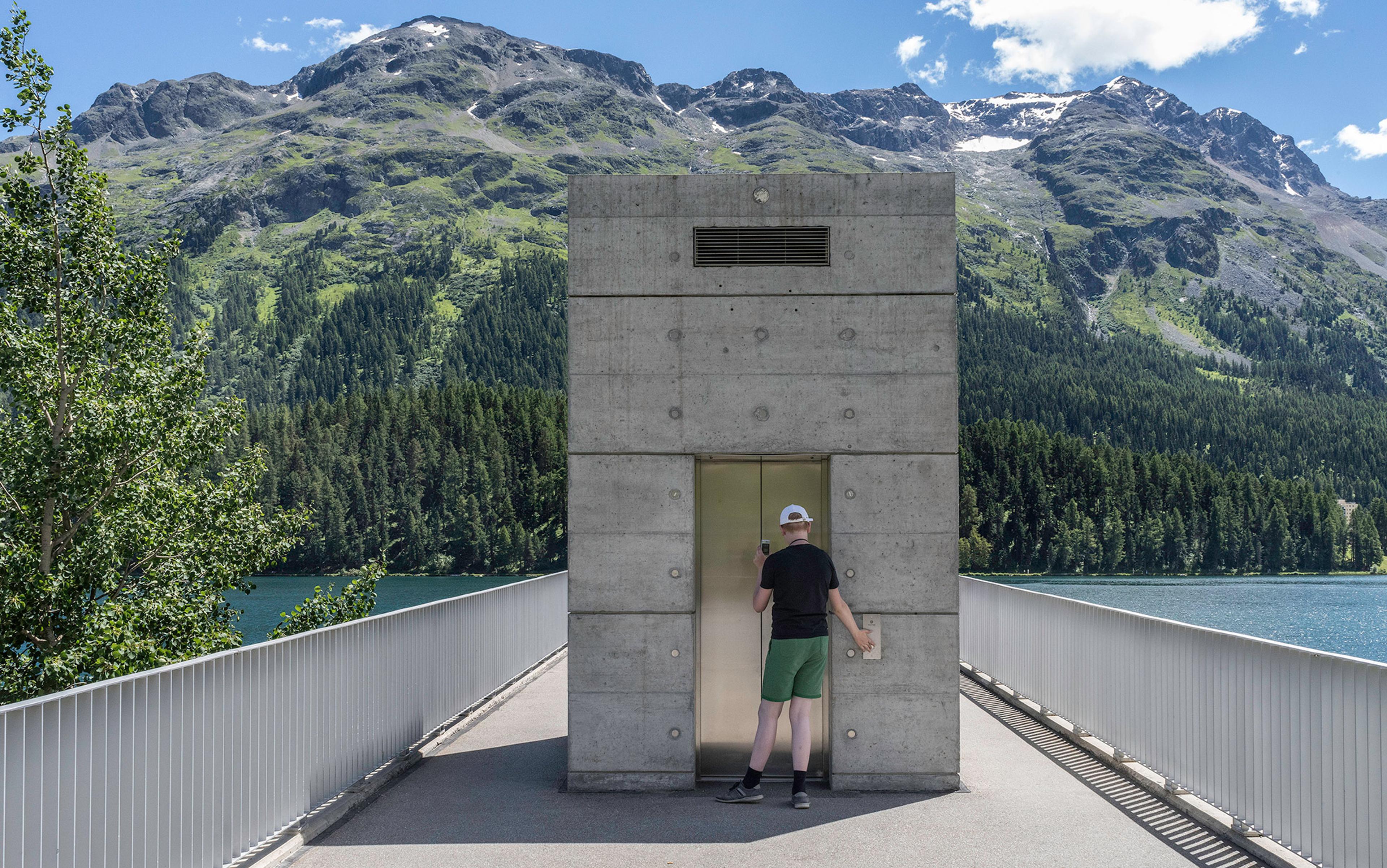 Photo of a person waiting by a concrete lift shaft on a bridge with trees, a lake and mountains in the background under a blue sky.