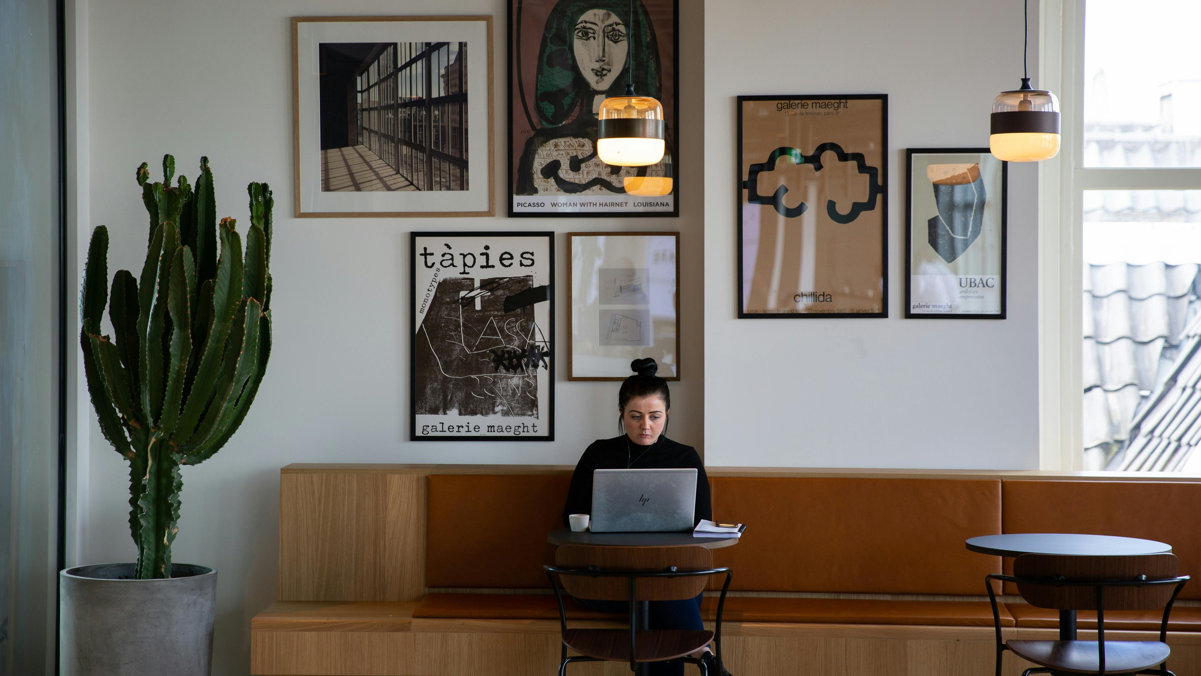 A woman on a laptop in a modern café with wall art, a large cactus and pendant lights above.