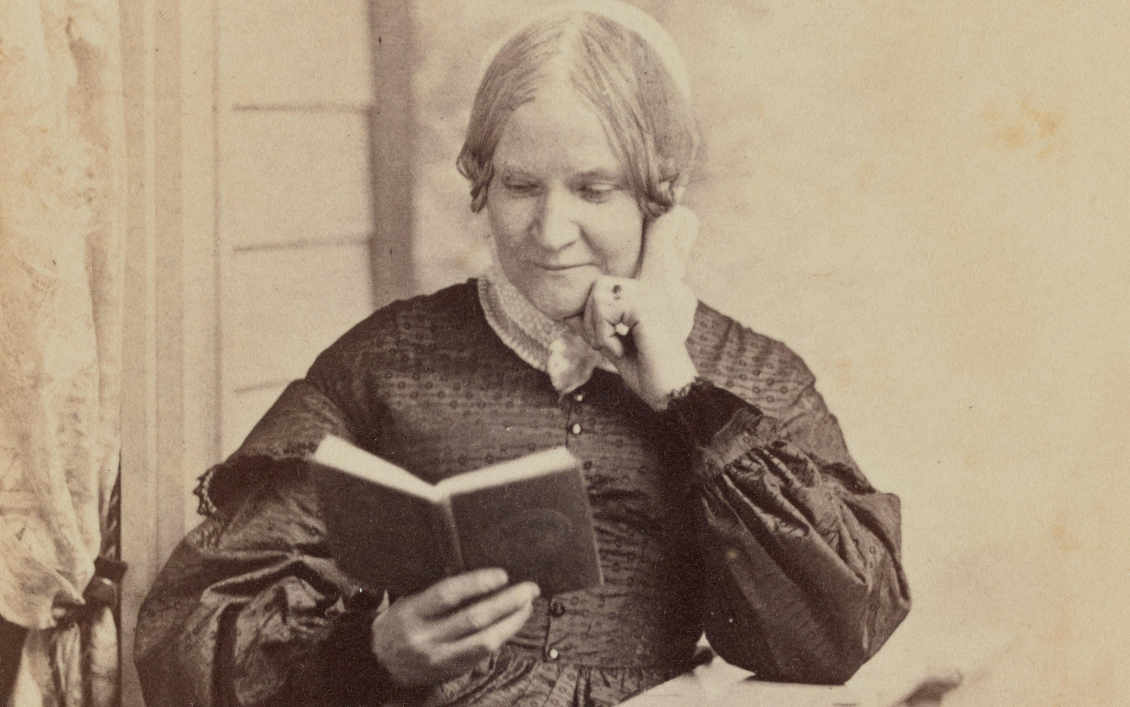 A sepia-toned, historical photograph of a woman sitting and reading a book, wearing a dark dress with puffed sleeves.