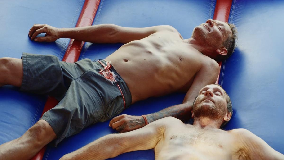 Photo of two shirtless men lying on a blue inflatable structure, eyes closed, one wearing shorts with patterned stripes.