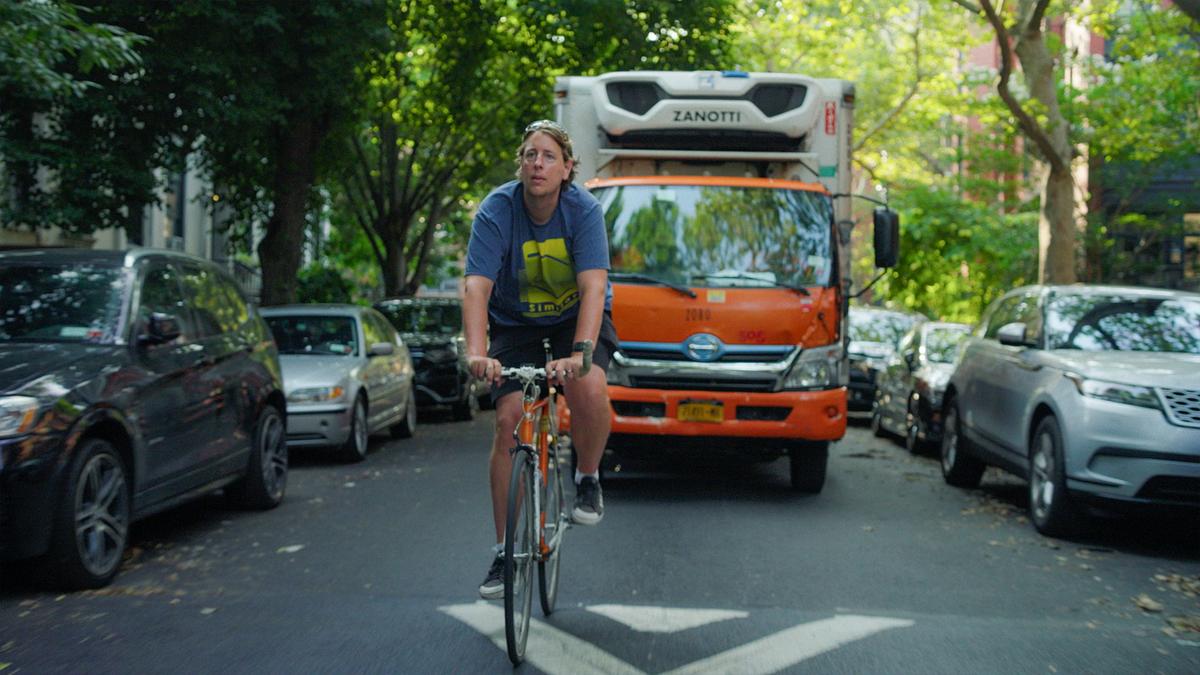 Photo of a person cycling on a city street with parked cars and an orange truck in the background under green trees.