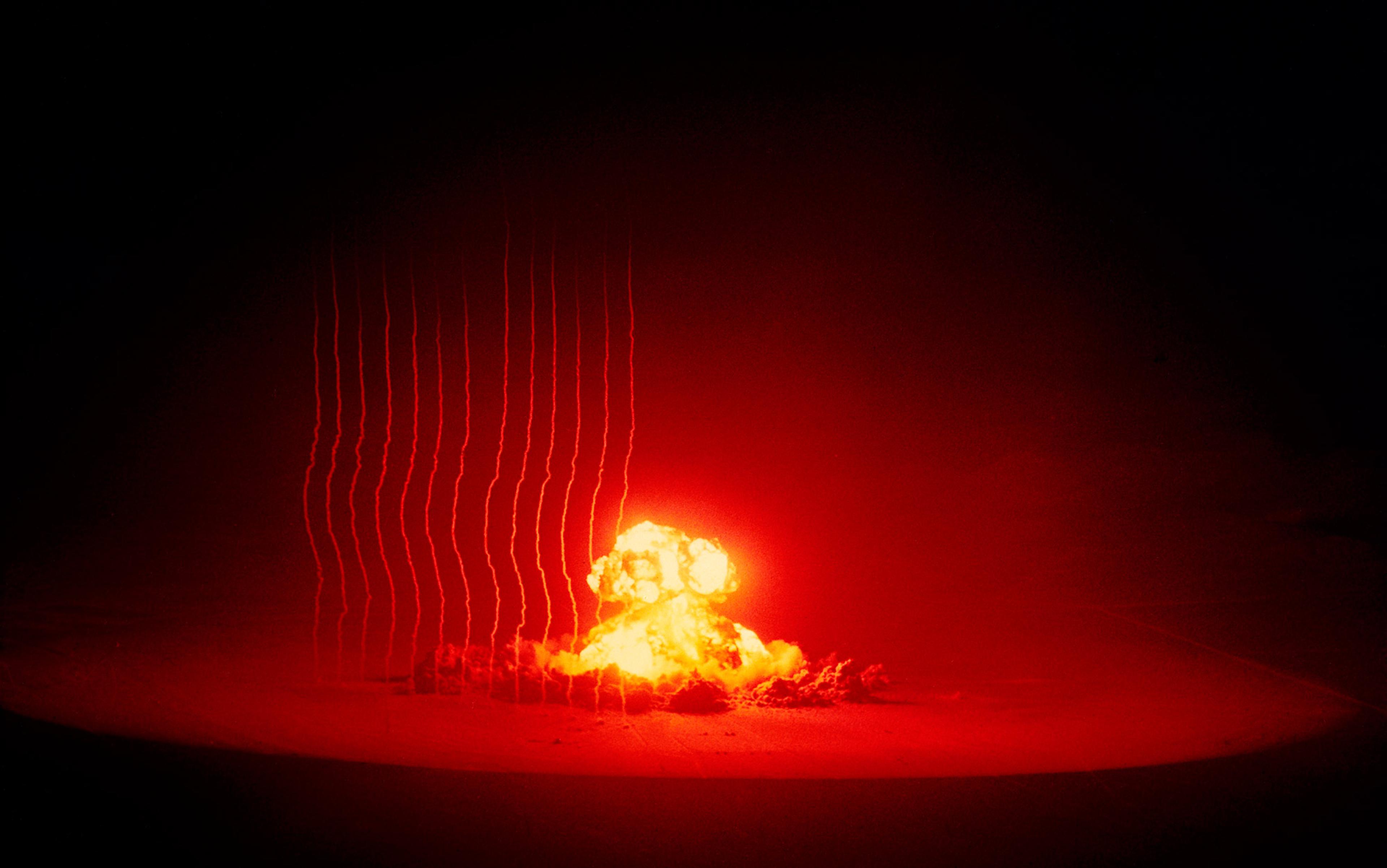 A nuclear explosion at night with bright red and yellow flames, a mushroom cloud and light trails on the left side.