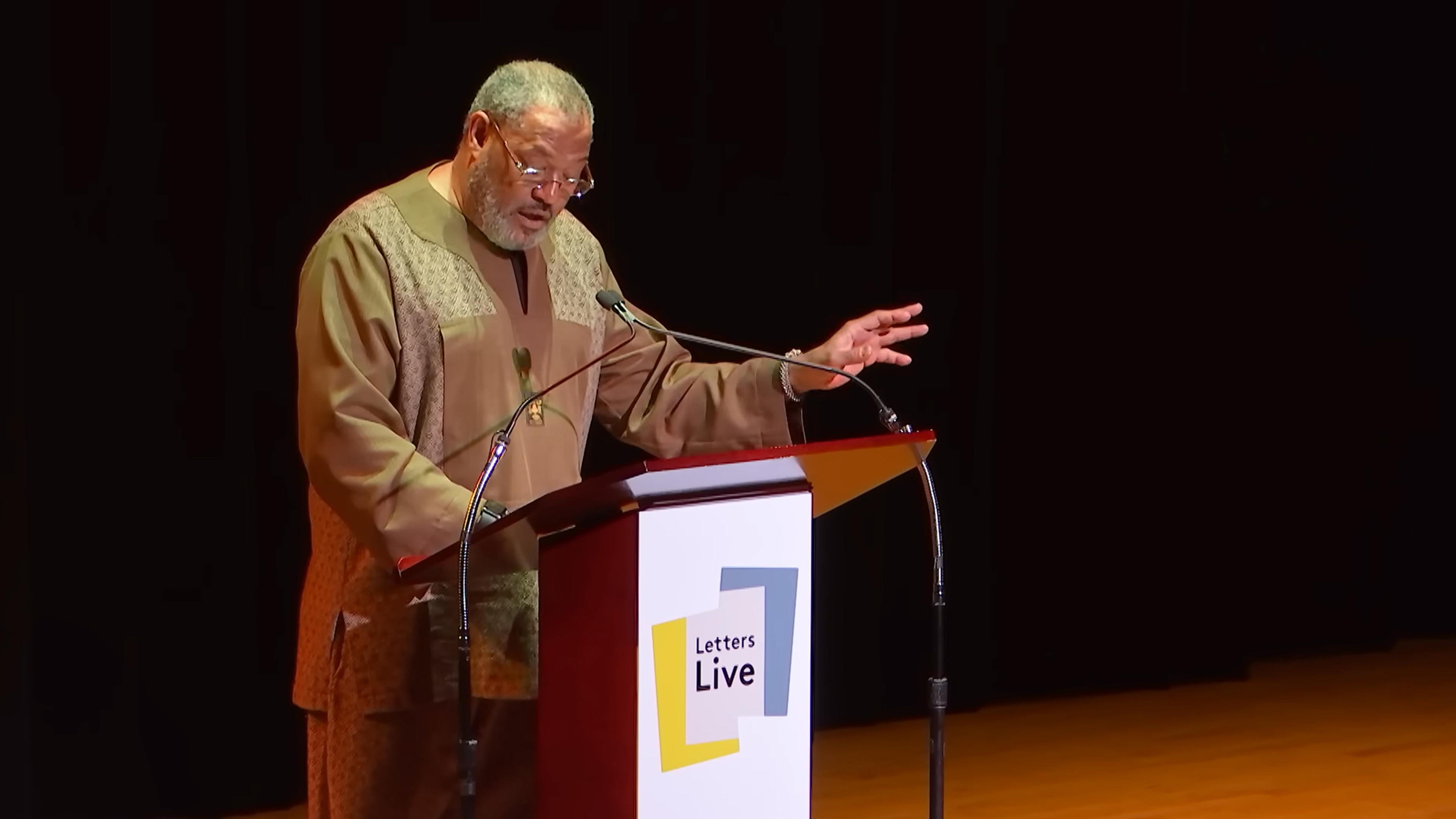 Photo of a man speaking at a podium marked ‘Letters Live’, gesturing with one hand against a dark background.