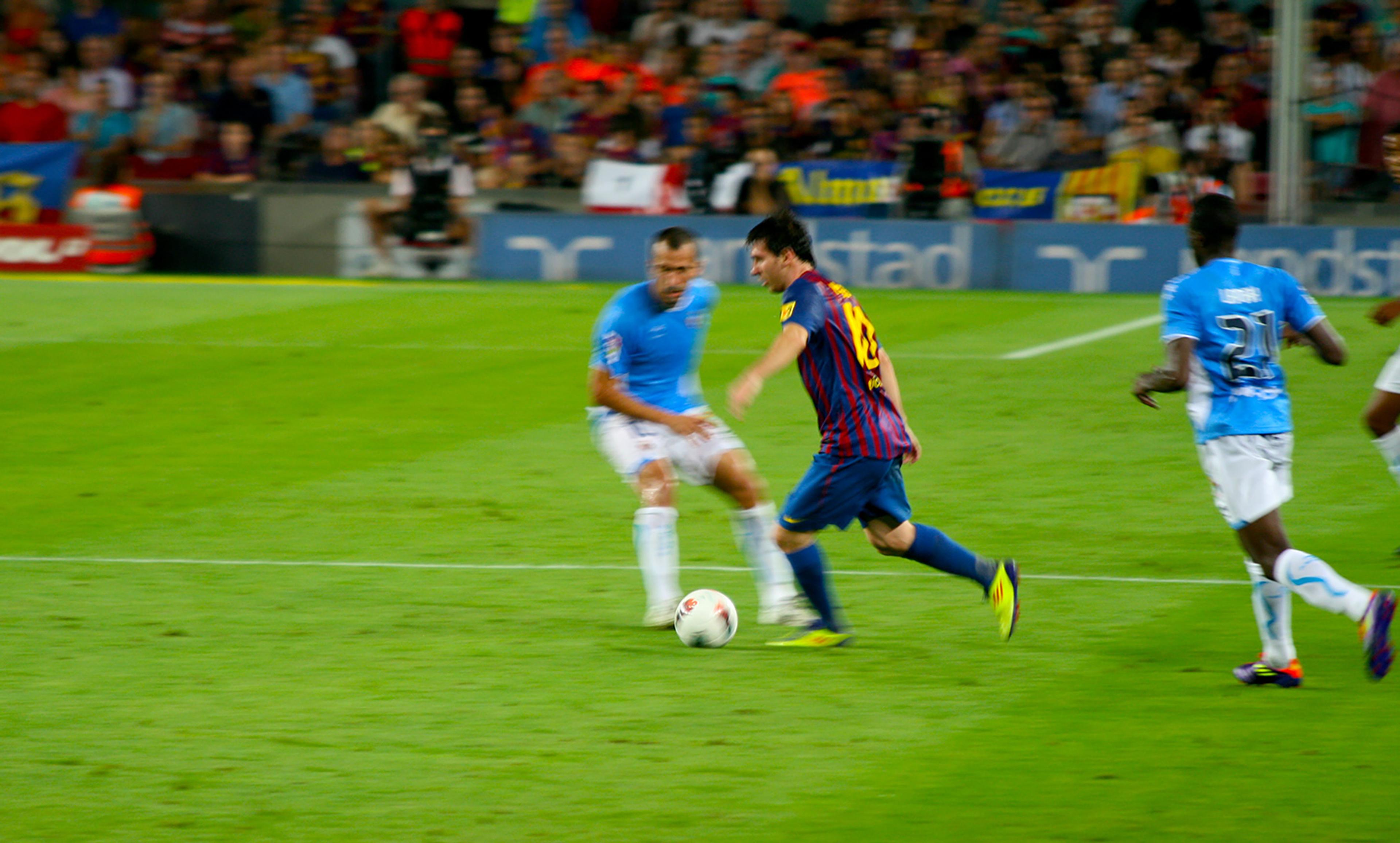 A football match with players in action, one in blue and white, another in red and blue, with a blurred crowd in the background.