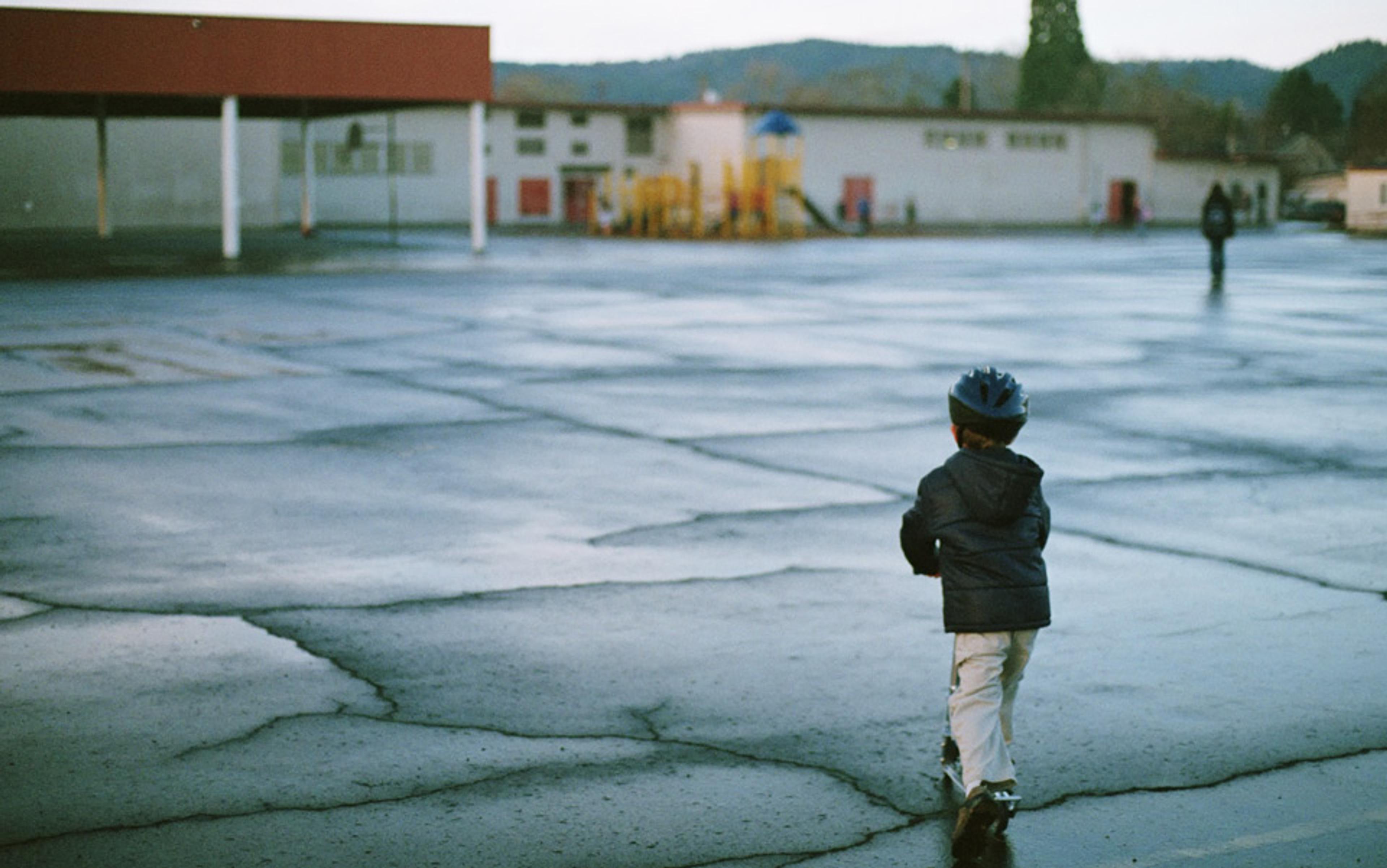 A child in a helmet riding a scooter in a wet, empty school playground with a building, playground equipment and hills in the background.
