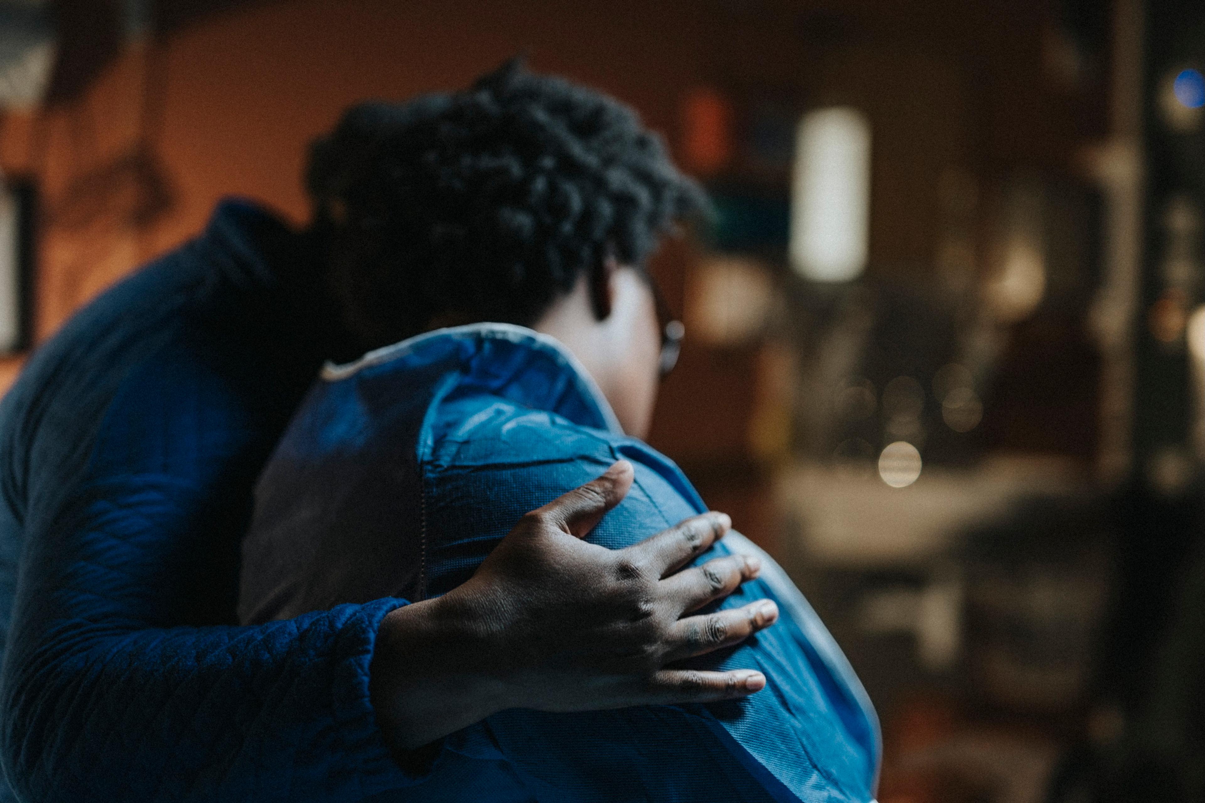Photo of two people embracing one another, with a hand gently placed on the back of a person wearing a blue jacket.