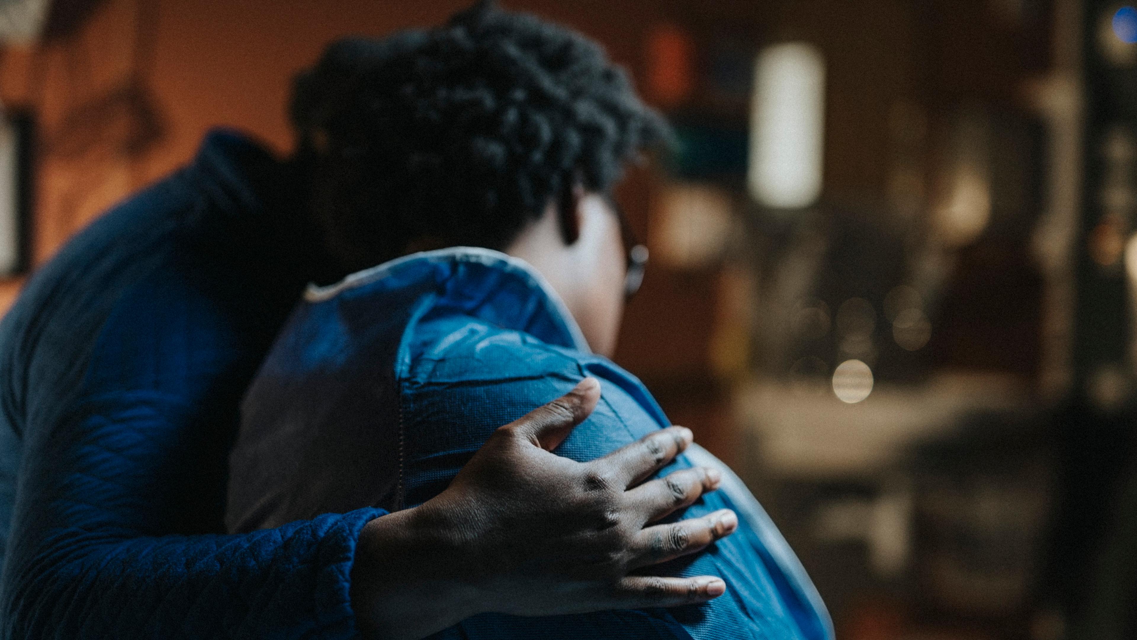 Photo of two people embracing one another, with a hand gently placed on the back of a person wearing a blue jacket.