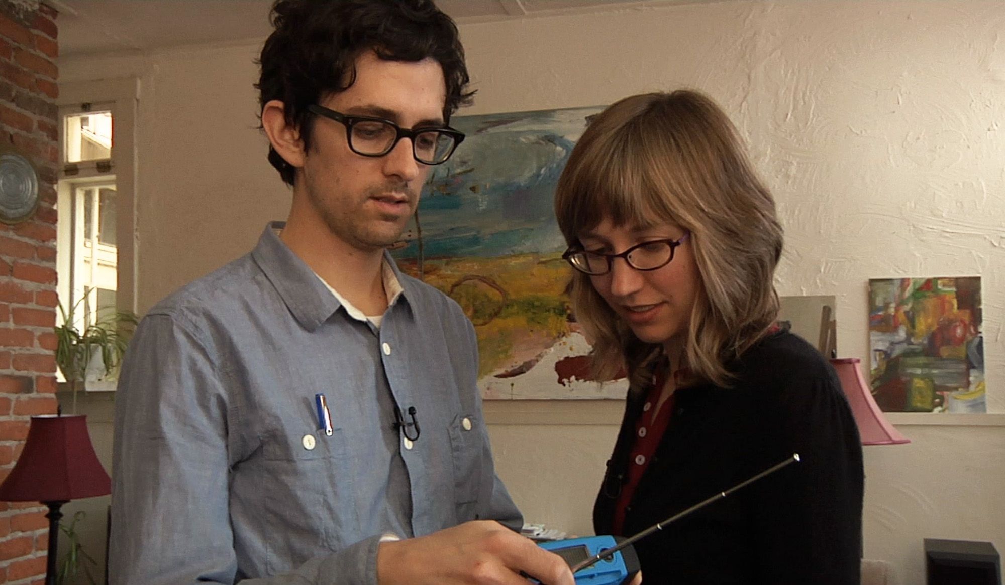 Photo of a man and woman with glasses examining a handheld device in an art-filled room with brick wall and paintings.