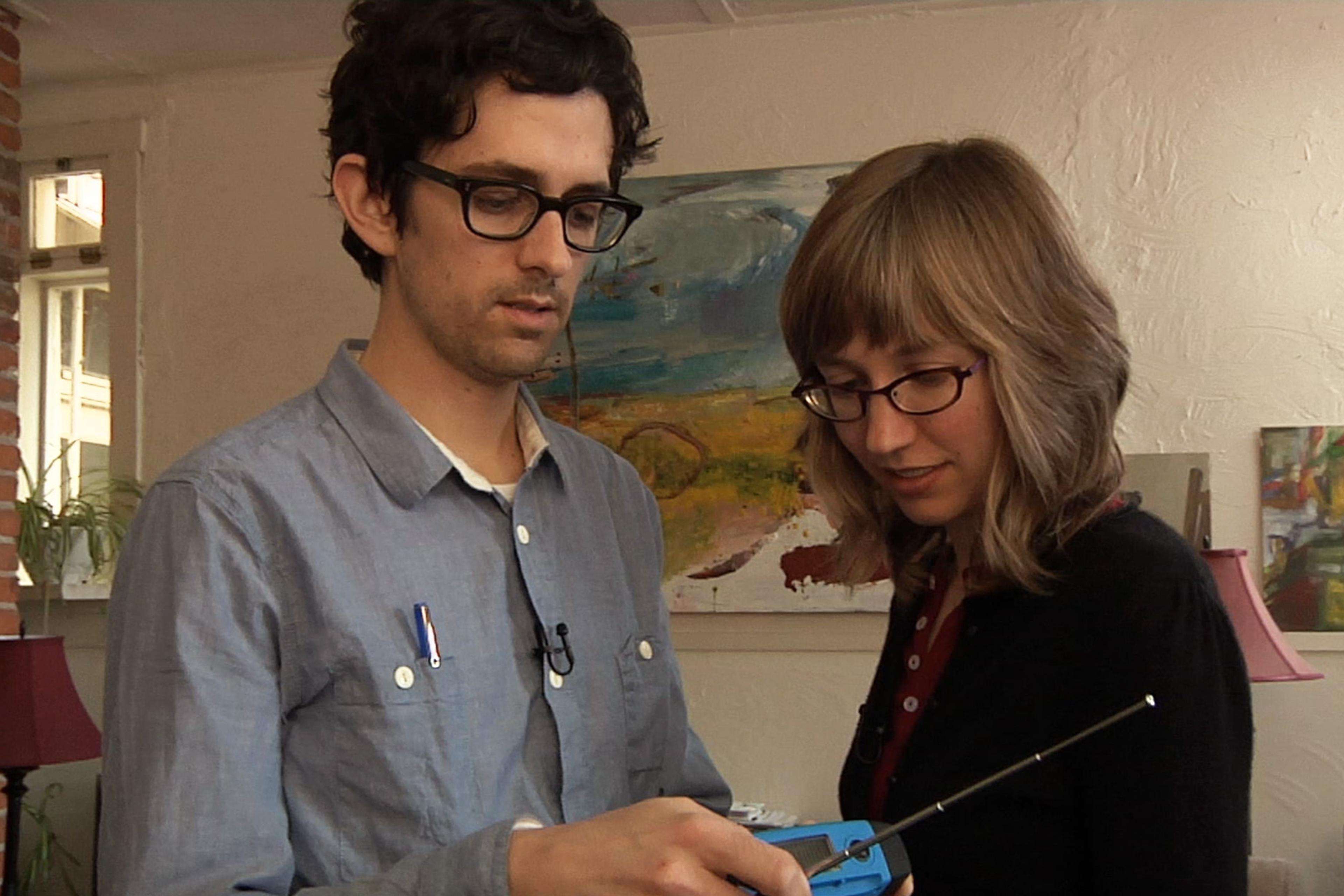Photo of a man and woman with glasses examining a handheld device in an art-filled room with brick wall and paintings.