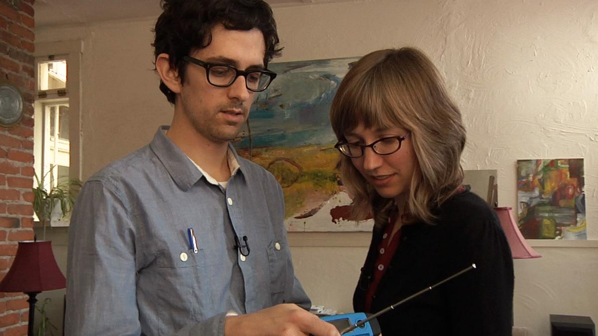 Photo of a man and woman with glasses examining a handheld device in an art-filled room with brick wall and paintings.