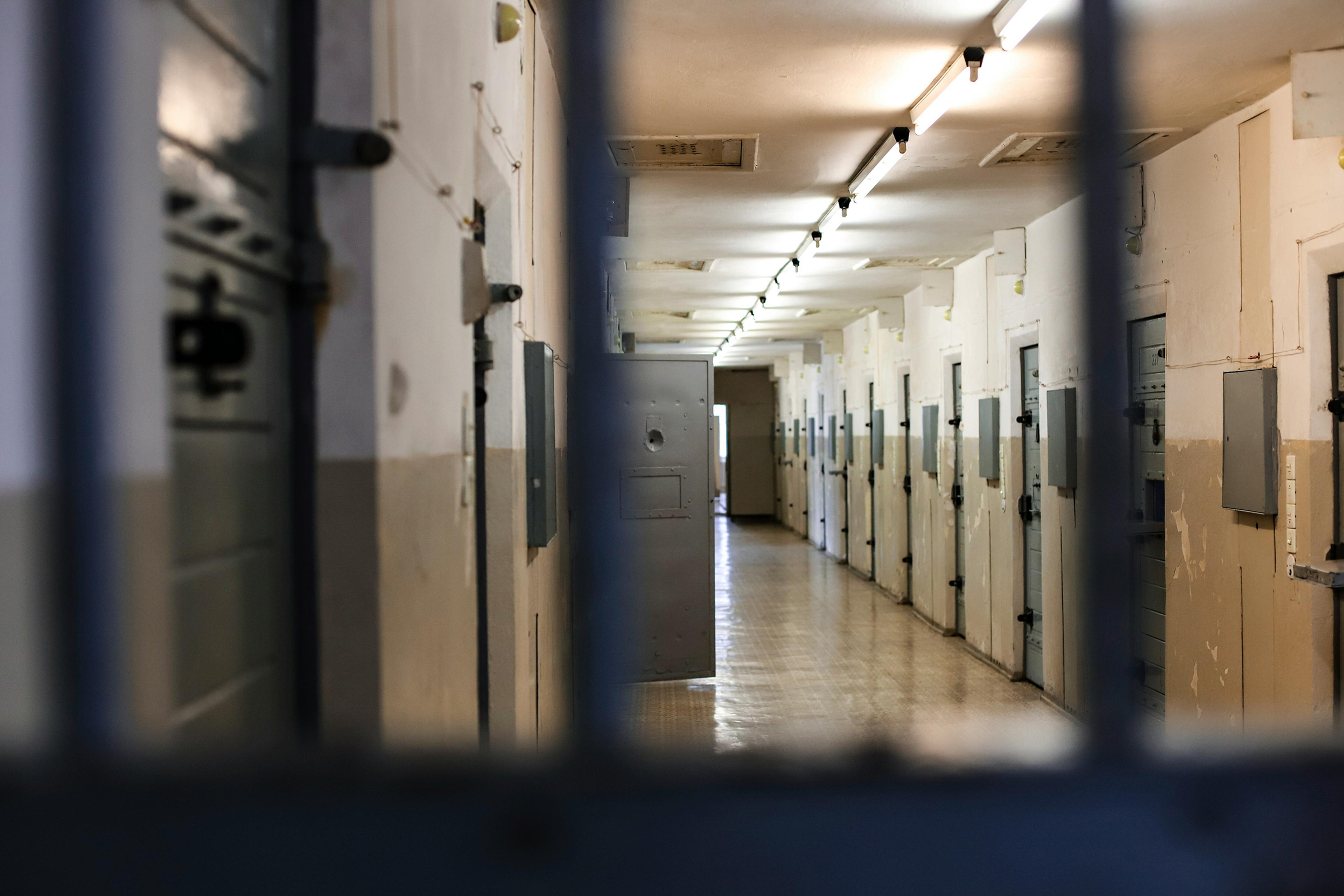 A dimly lit prison corridor with closed cell doors on both sides viewed through metal bars.