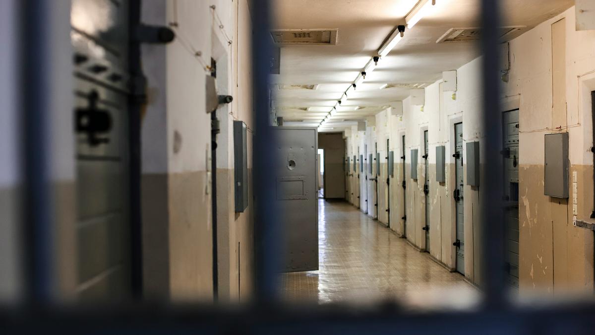 A dimly lit prison corridor with closed cell doors on both sides viewed through metal bars.