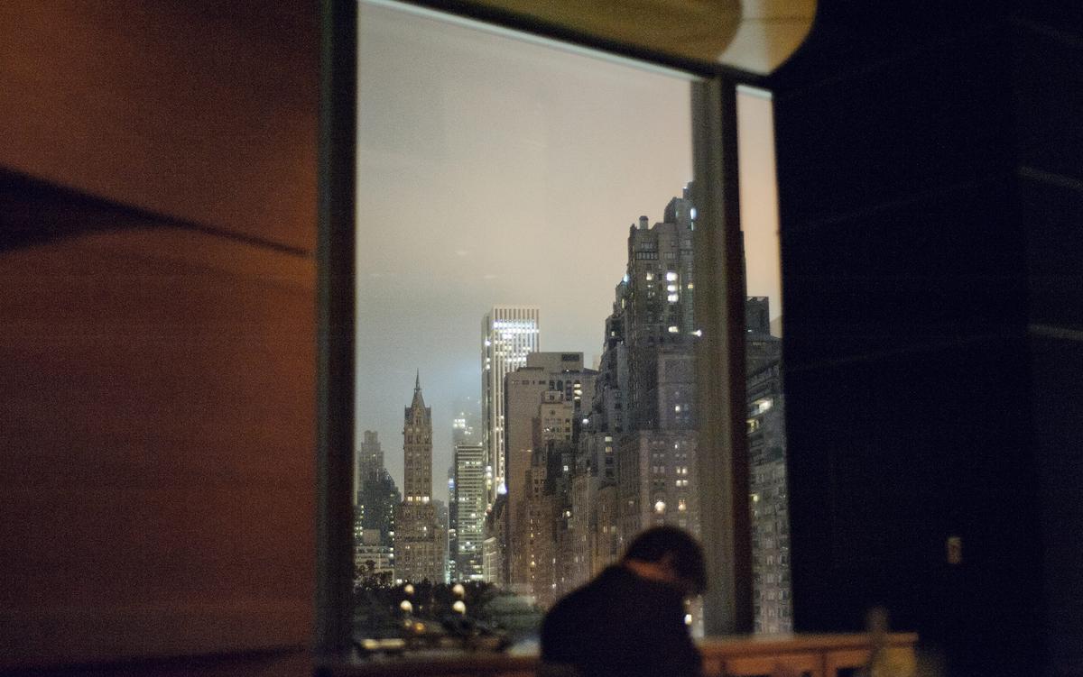 A cityscape at night through a window with a person in the foreground, blurred buildings in the background.