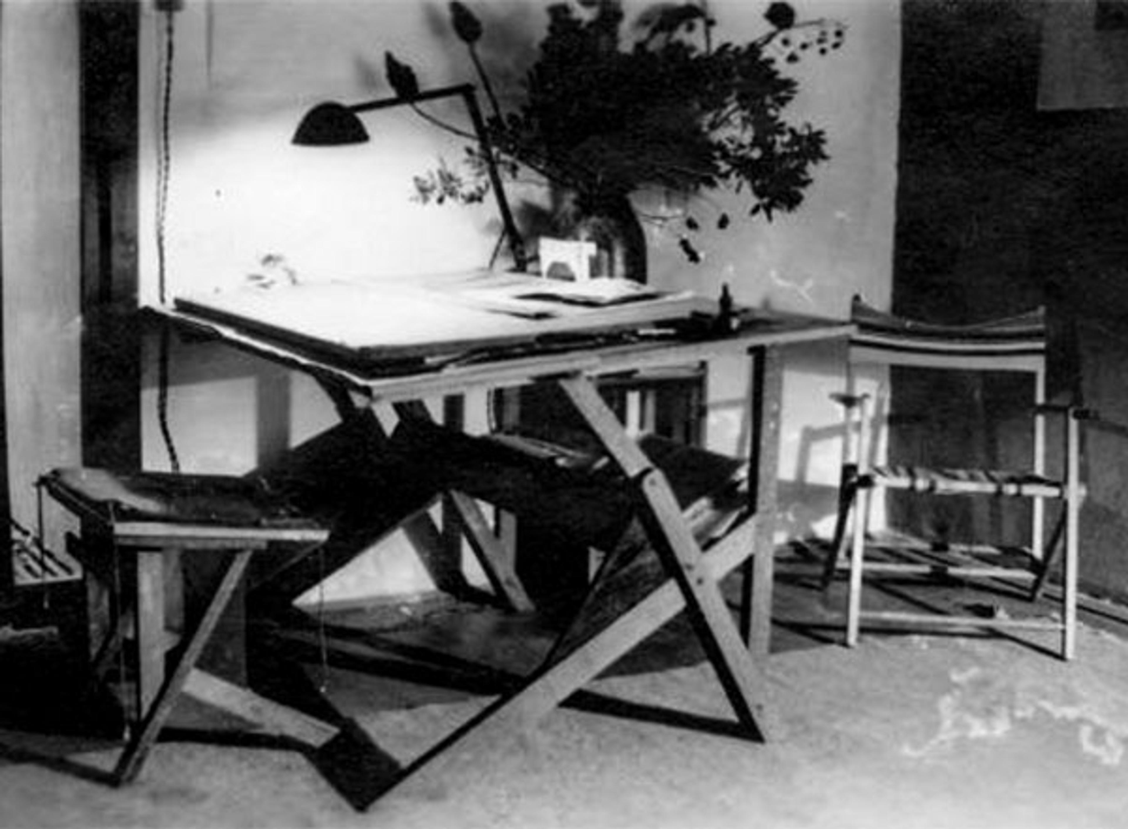 Black and white photo of a drafting table with a stool a lamp and a director’s chair in a room with a plant in the background.