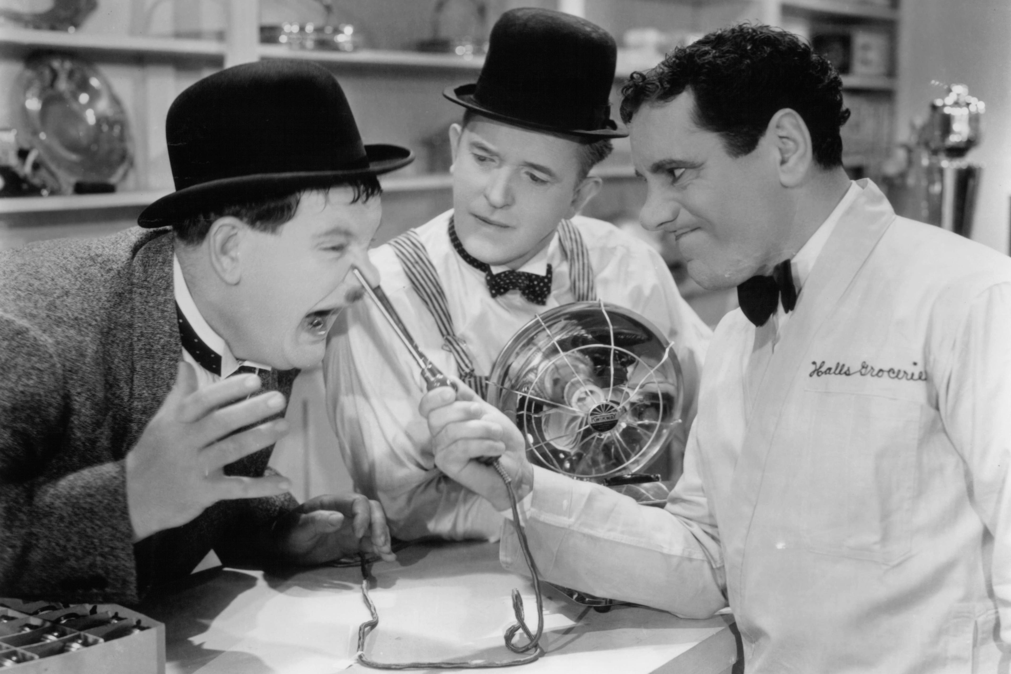 Black and white photo of three men in a shop, two in bowler hats clowning with an electric fan while a clerk watches amusedly.