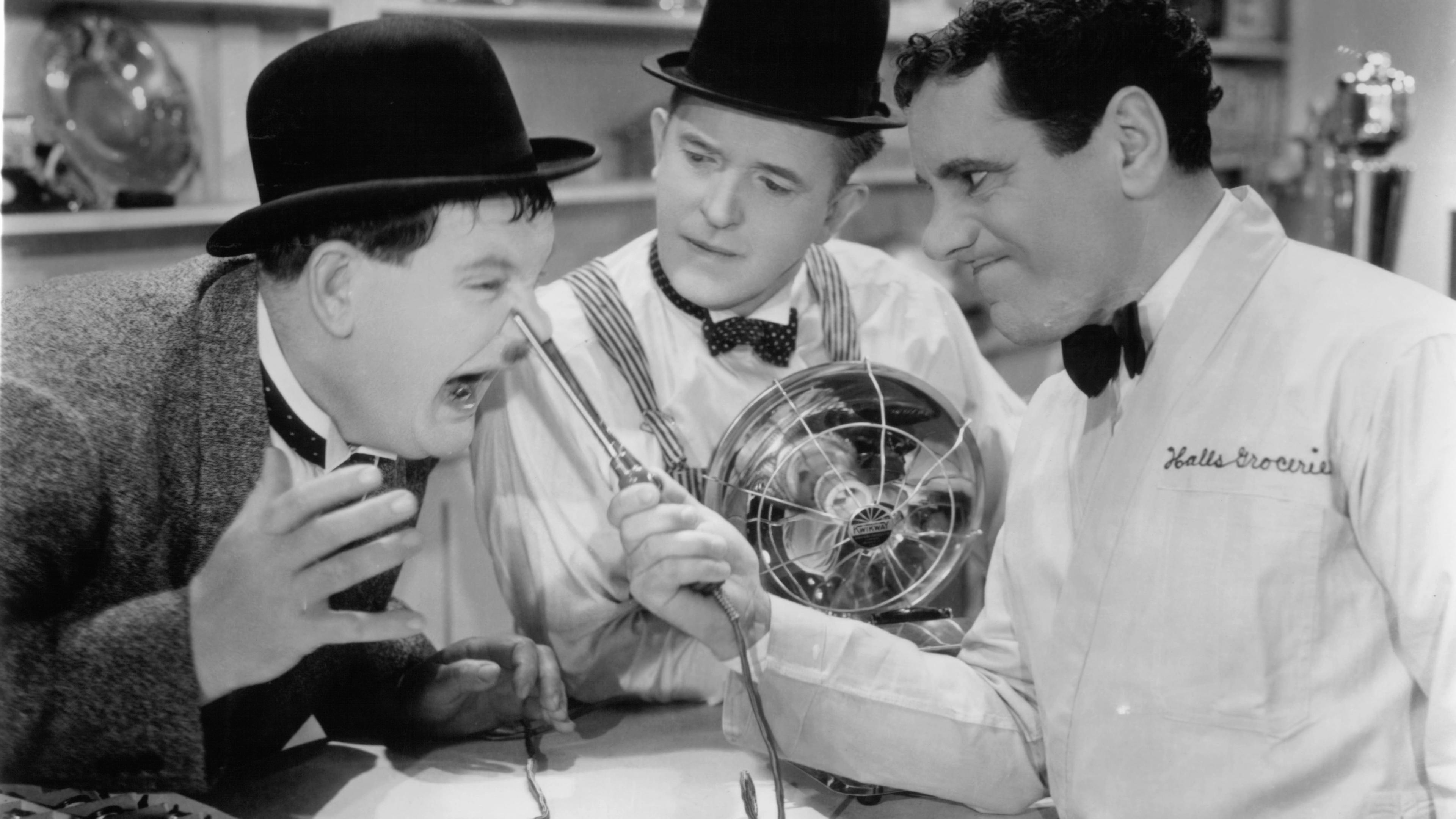 Black and white photo of three men in a shop, two in bowler hats clowning with an electric fan while a clerk watches amusedly.