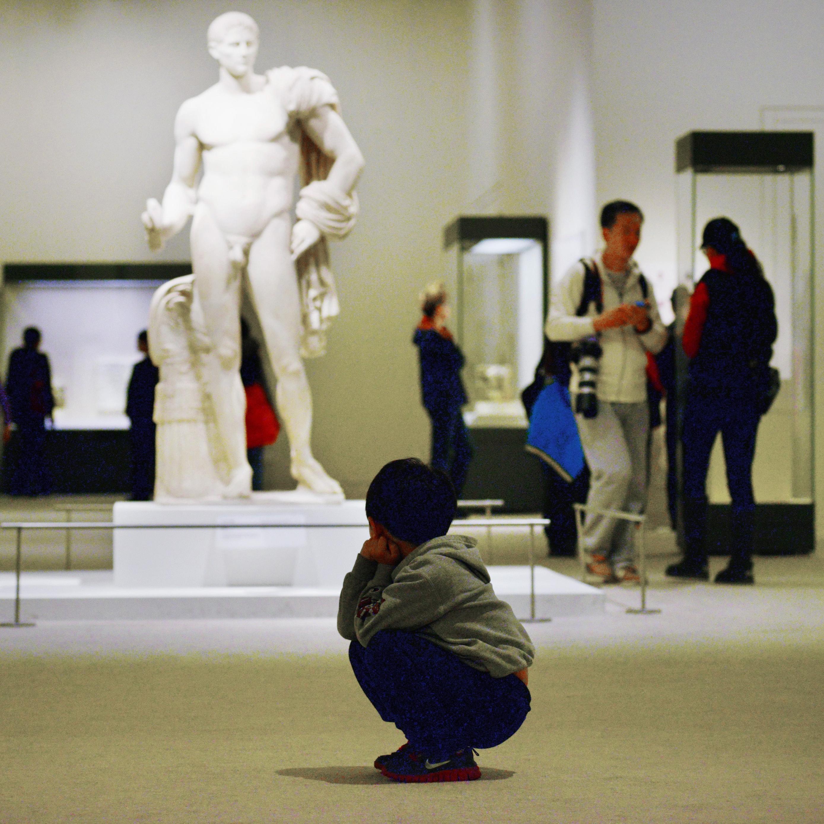 Photo of a child squatting in a museum, observing a large classical statue with people around.