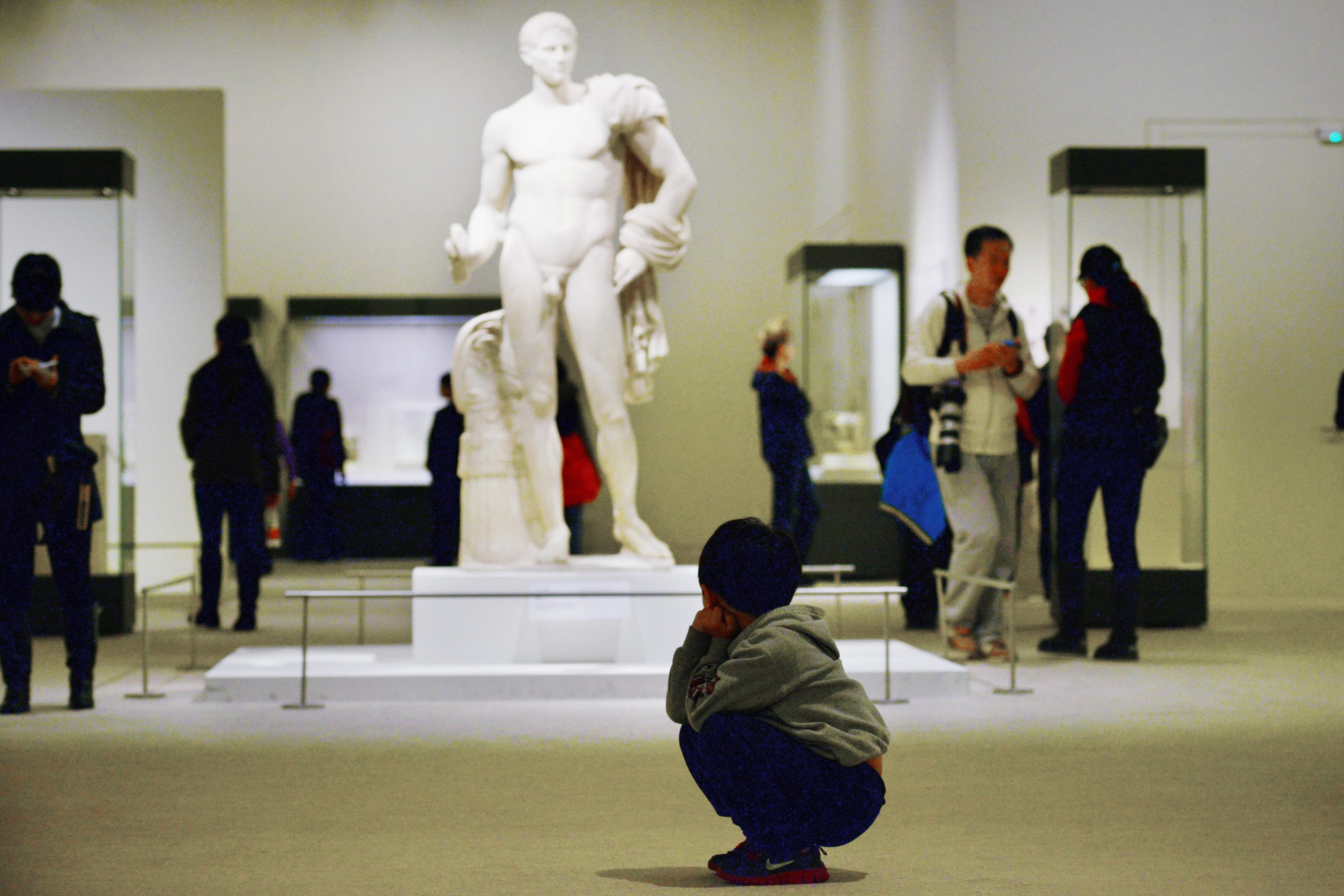 Photo of a child squatting in a museum, observing a large classical statue with people around.